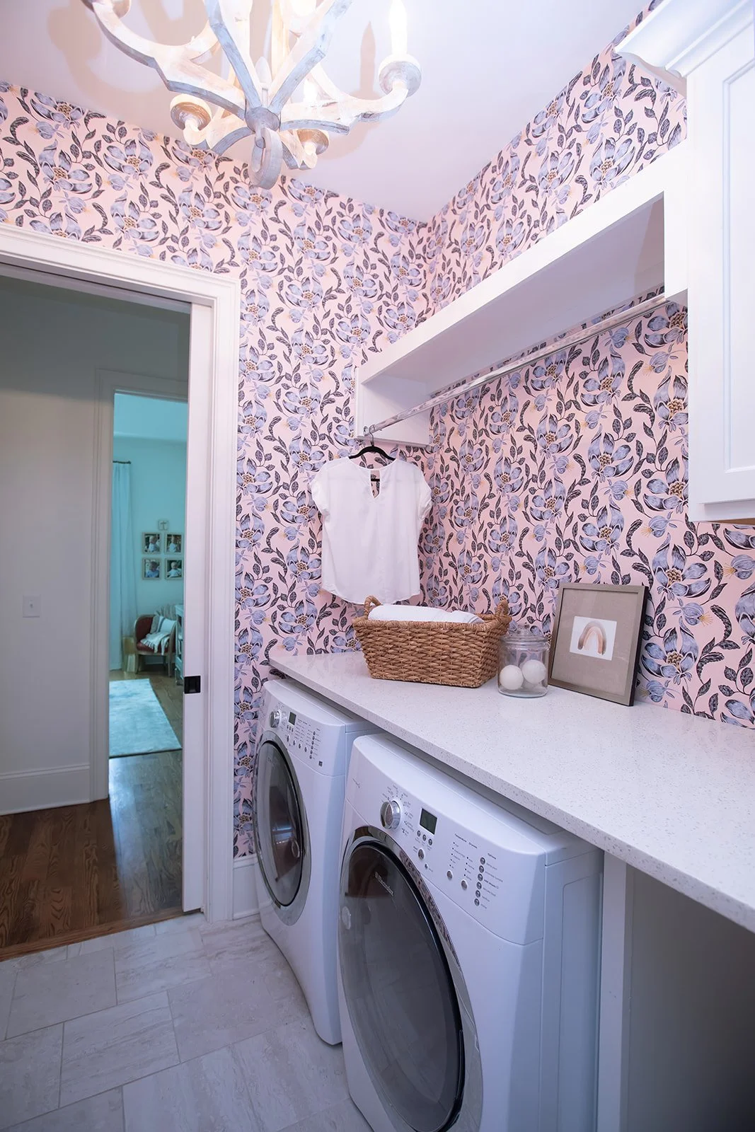 Laundry room with floral wallpaper, featuring white front-loading washer and dryer, white shelves, wicker basket, and framed picture.