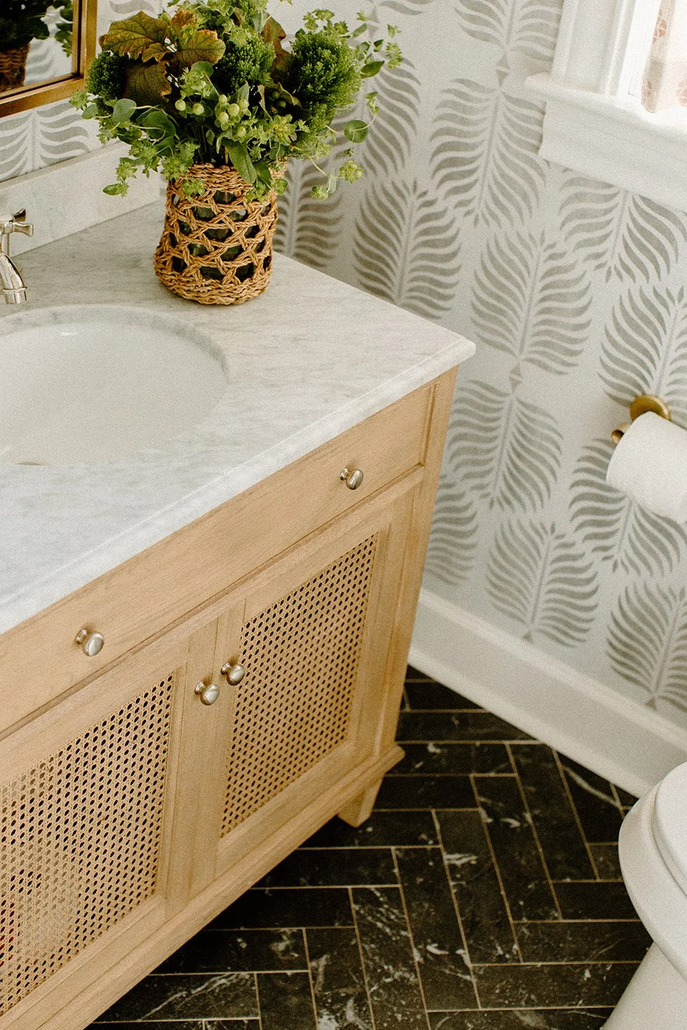Bathroom vanity with marble countertop, wicker cabinet, potted plant, leafy wallpaper, and black herringbone tile floor.