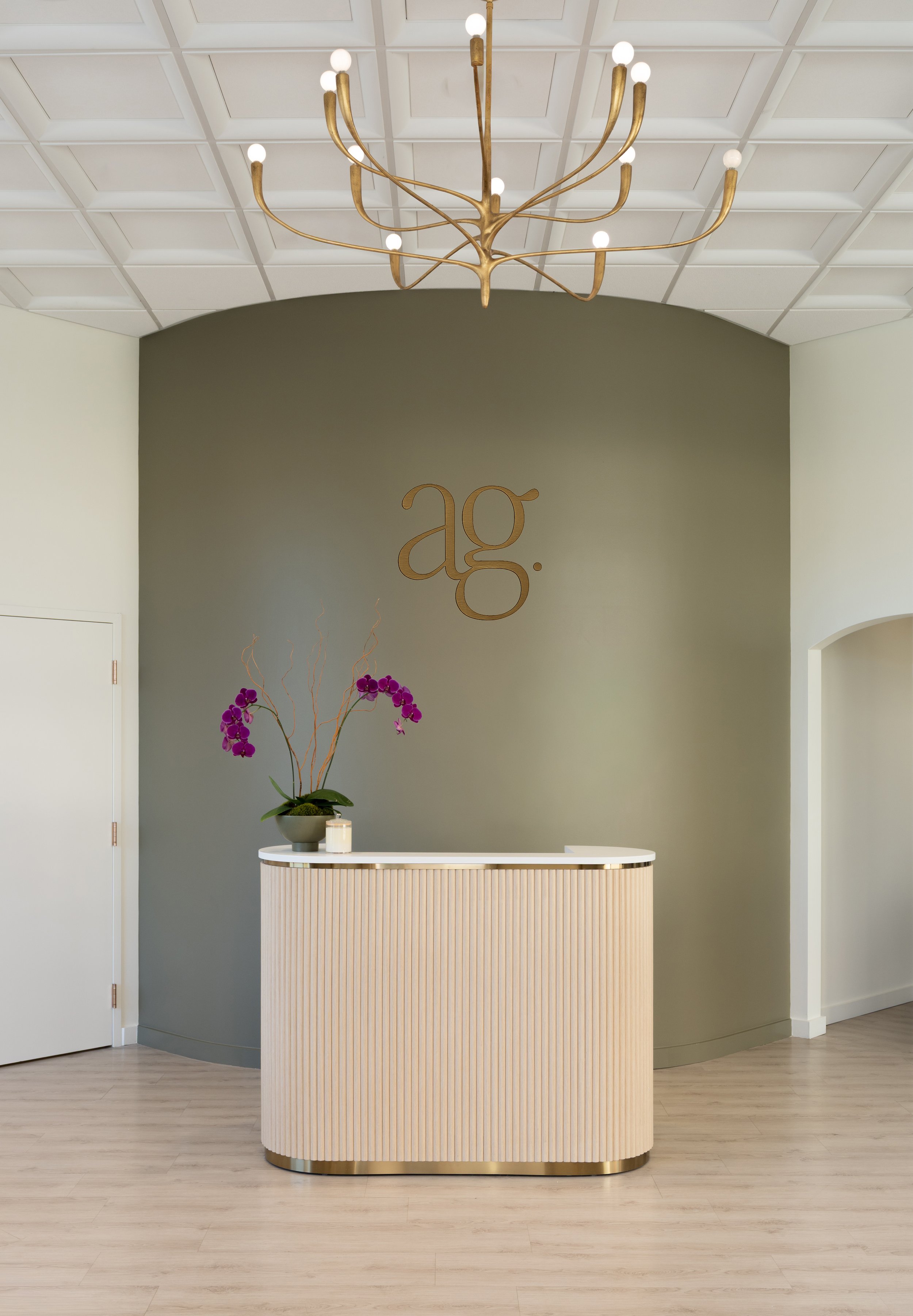 Modern reception desk with purple orchids on top, a green wall with gold "ag." logo, white ceiling tiles, and a gold chandelier.
