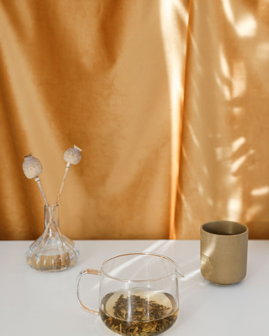 Tasse en verre avec infusion de thé, vase en verre avec deux tiges florales séchées, et un gobelet en métal sur une surface blanche devant un rideau en tissu doré