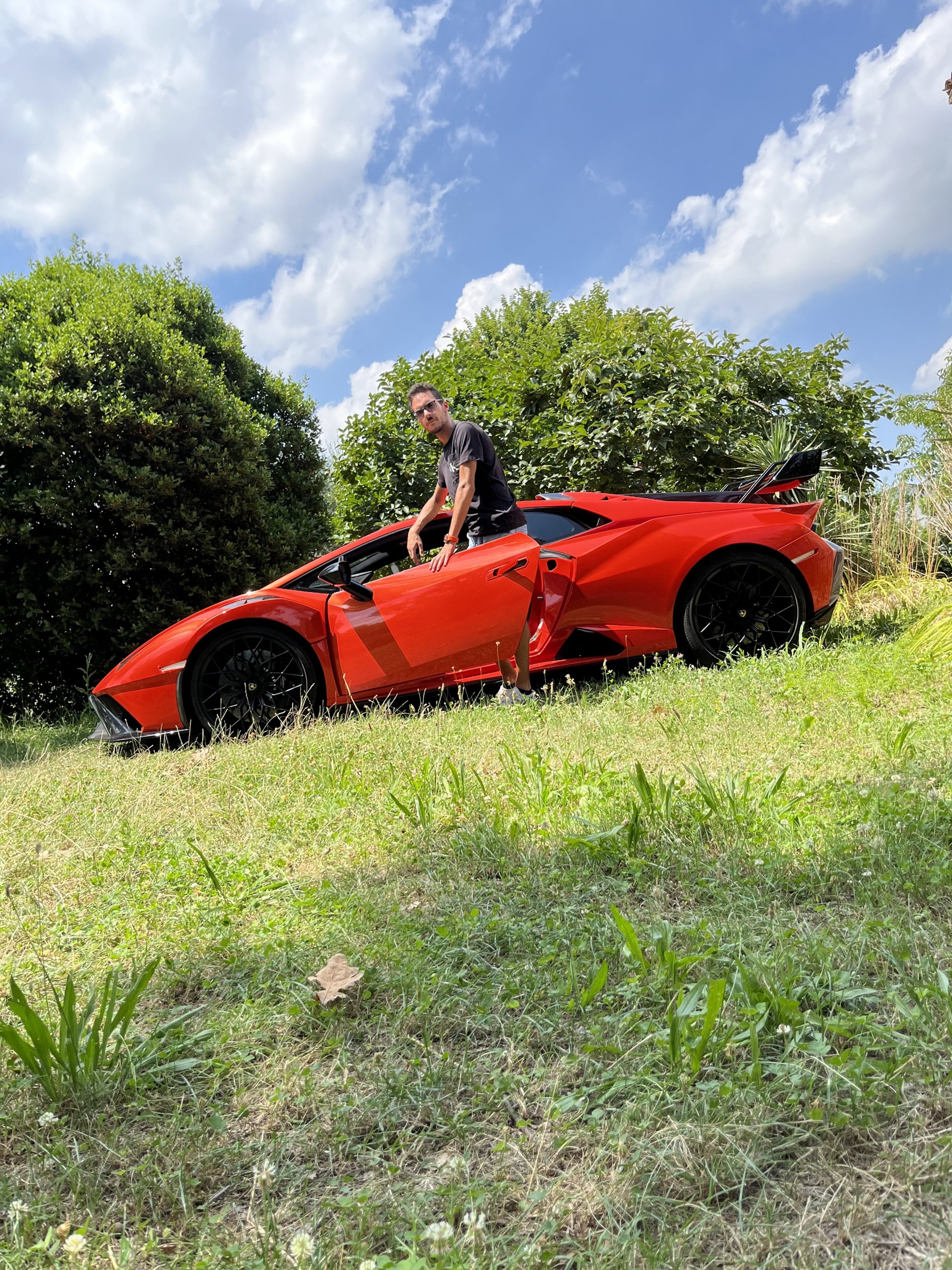 Uomo accanto a un'auto sportiva rossa in un prato, con alberi e cielo nuvoloso sullo sfondo.