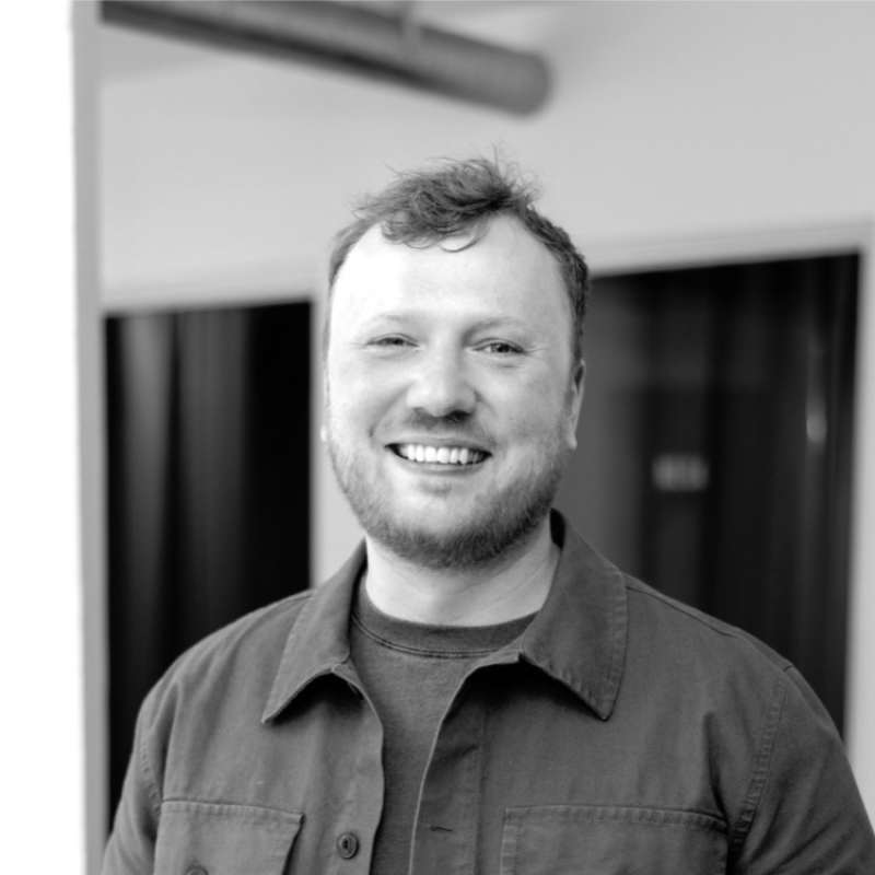 Black and white photo of a smiling man with a beard and short hair, wearing a dark shirt, in an indoor setting with curtains and wall in the background.