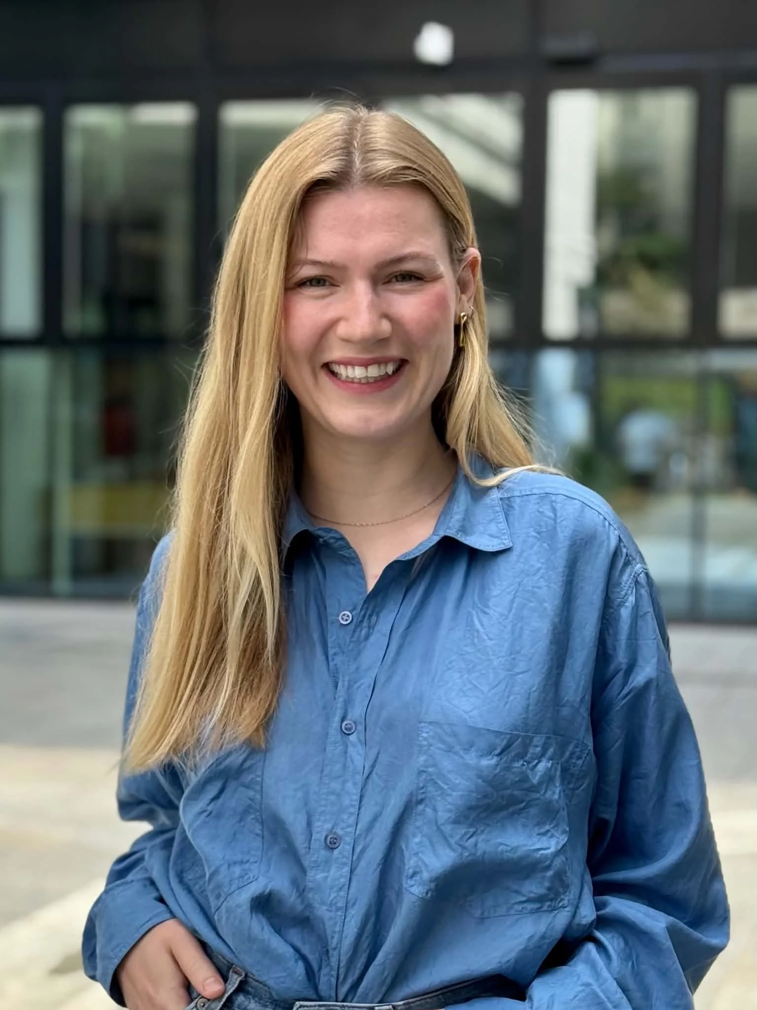 Et smilende kvinne med langt blond hår og blå skjorte som sitter utendørs foran et moderne bygg med glassfasade.