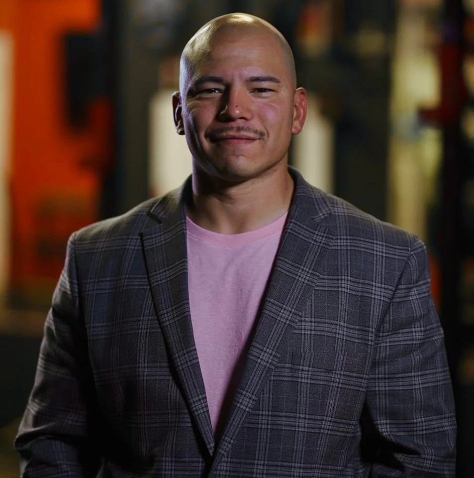 Jessie Casas, Owner of Bold Strength Fitness, wearing a plaid blazer over a pink shirt, standing indoors with a blurred background.