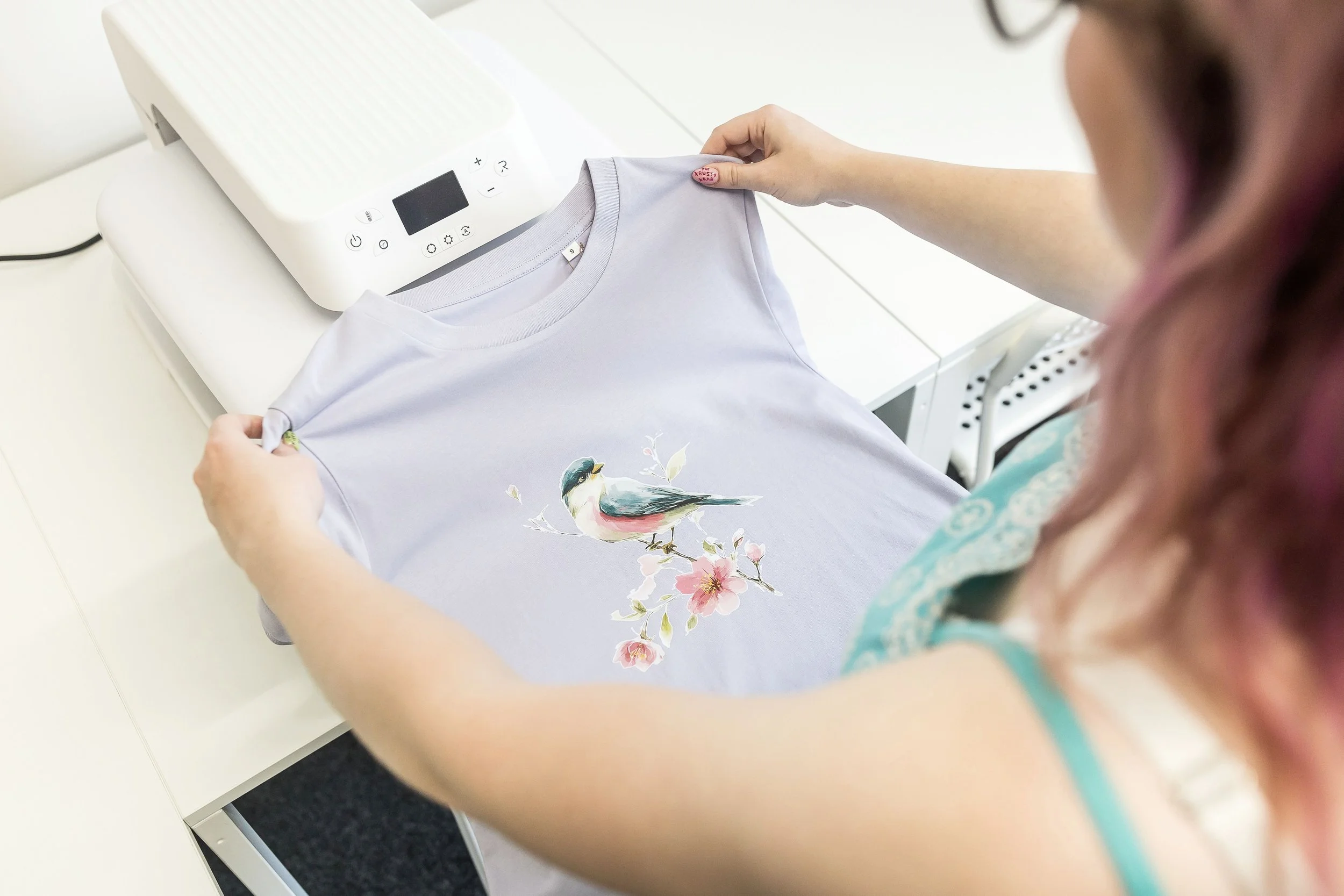Freshly printed lilac T-shirt with a watercolour bird design next to a heat press