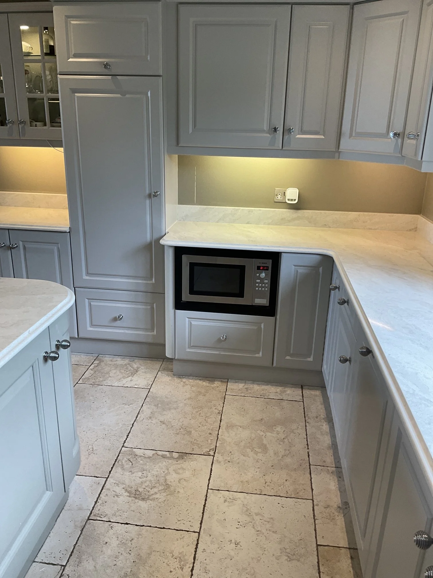 View of a kitchen with white cabinetry, a microwave built into lower cabinets, a beige countertop, and beige tile flooring with visible grout lines.