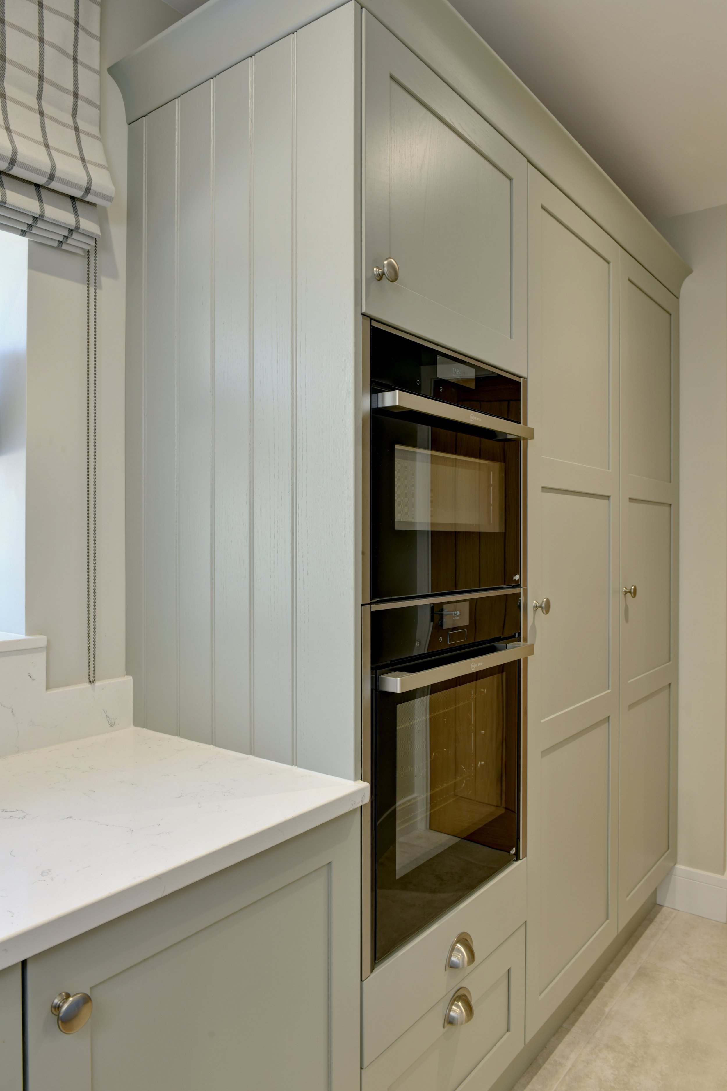 Kitchen corner showing built-in oven, greige cabinets, and a white marble countertop, with a window with checkered curtains.