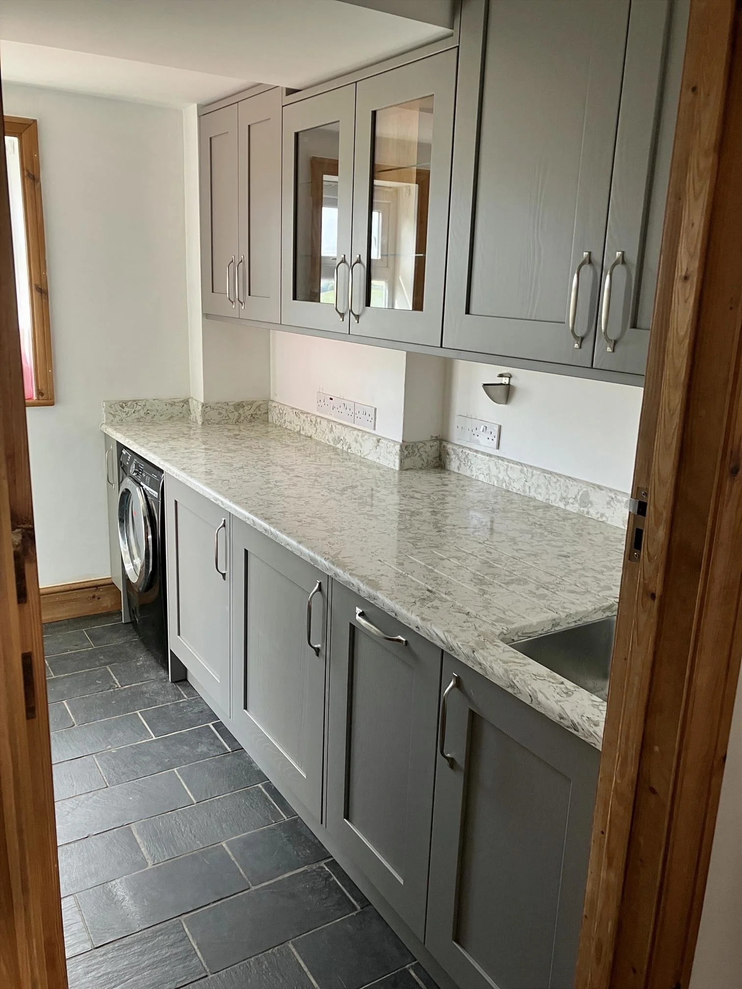 A modern utility room with grey cabinets, a marble countertop, a built-in sink, and a washing machine underneath the counter.