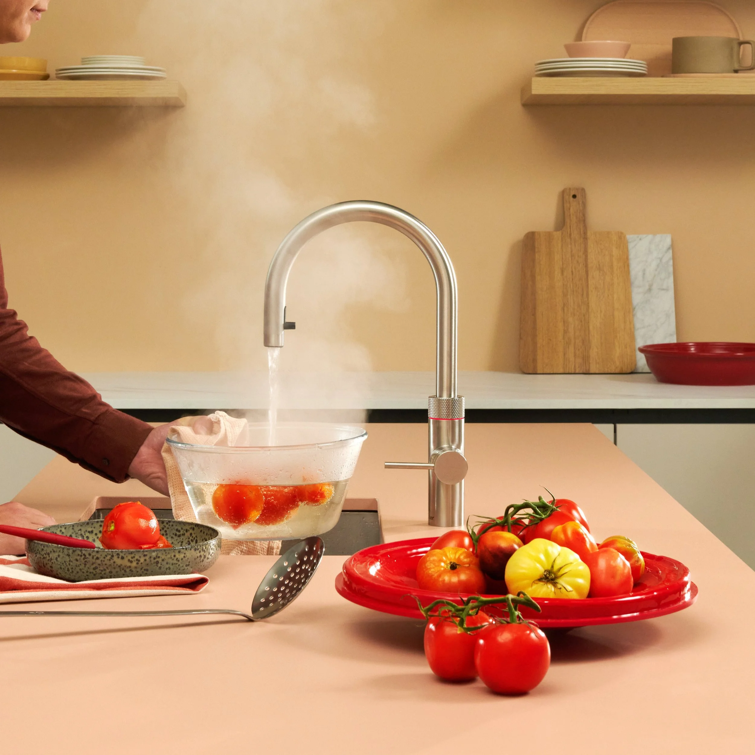 Quooker tap shown in hot mode. Person holding a glass bowl with water and tomatoes on a kitchen counter, with tomatoes on a red plate and cutting boards in the background.