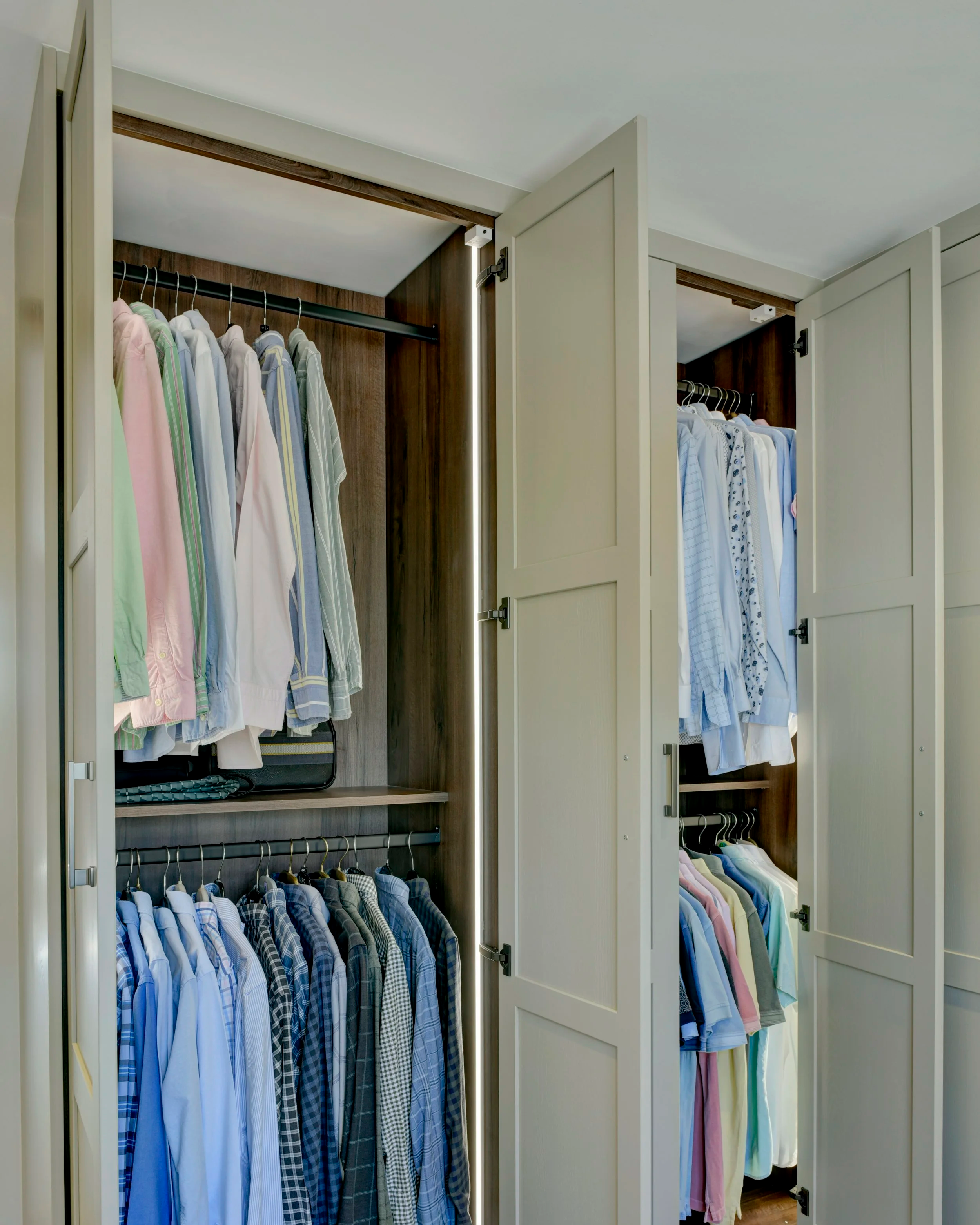 Open wardrobe with shirts hanging on racks, organised by color and pattern, with cream doors and wooden interior.