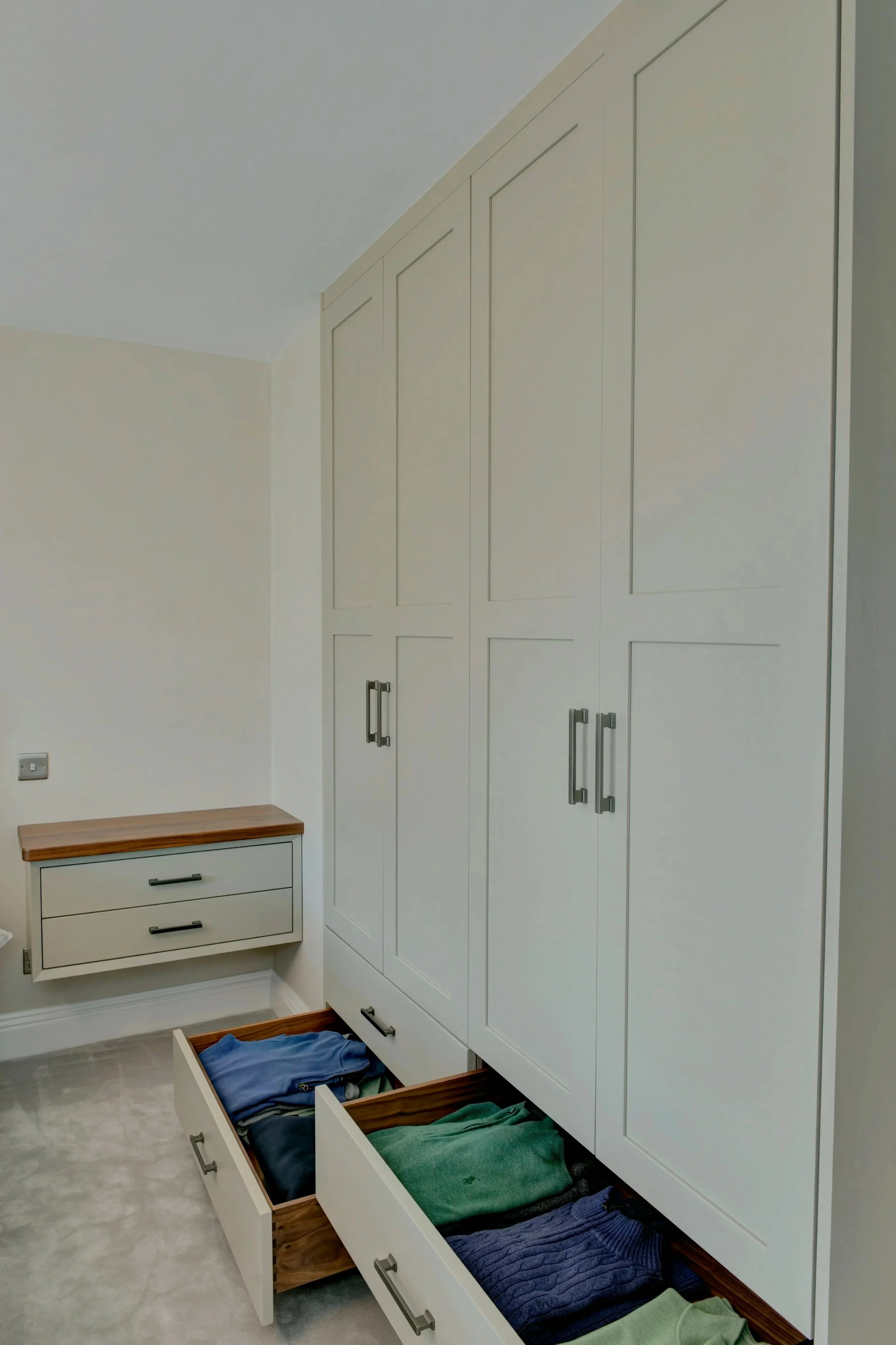 A bedroom with built in wardrobes showing an open drawer with folded clothes, and set of floating bed side drawers with a wooden top against a white wall. 