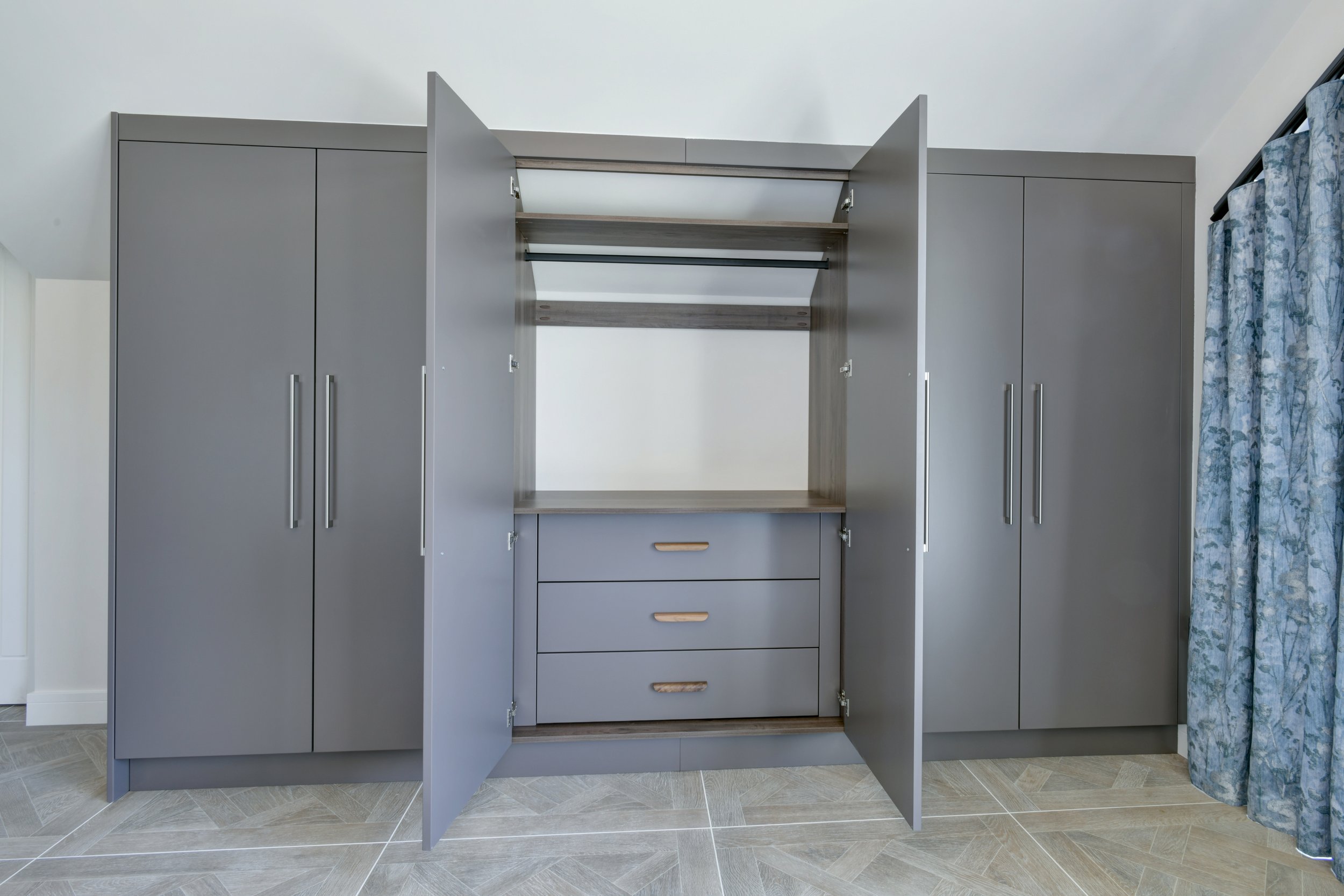 Built-in wardrobe with open doors revealing shelves and drawers, situated in a room with light-colored flooring and floral curtains.