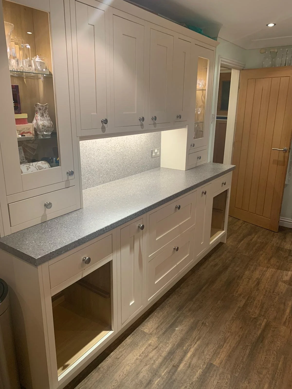 White kitchen cabinet with grey granite countertop and glass-door display sections, wooden door to the right, and wood flooring.