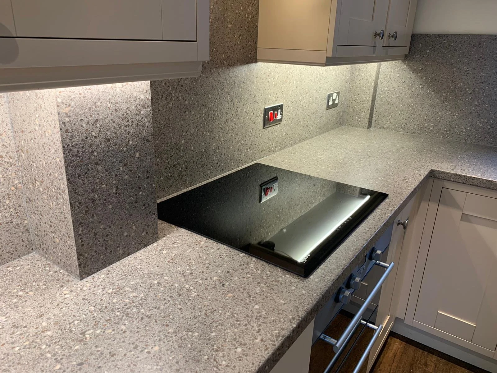 A kitchen countertop with an hob, grey cabinets and a speckled granite surface.