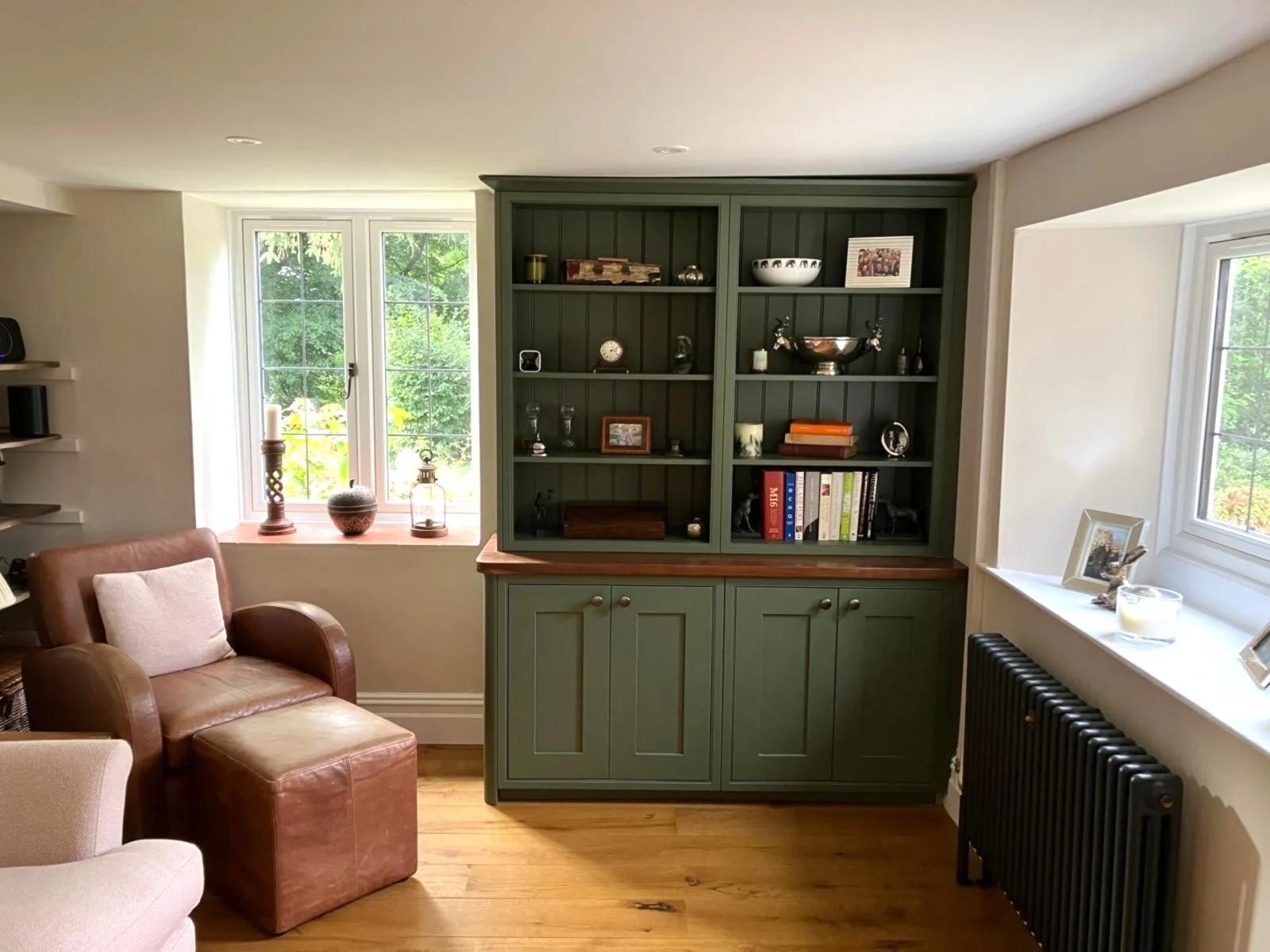 Living room with green wooden bespoke bookshelf, brown leather armchair, window sill with vases and lanterns, white walls, wooden floor, black radiator, and windows with greenery outside.