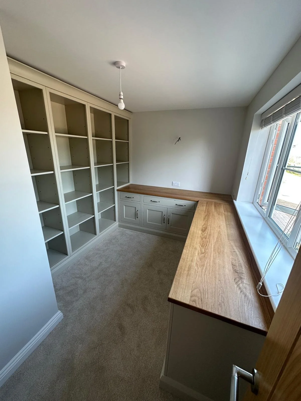 Office with built-in desk and shelves, a wooden countertop, grey cabinets, a large window with blinds, and beige carpet flooring.