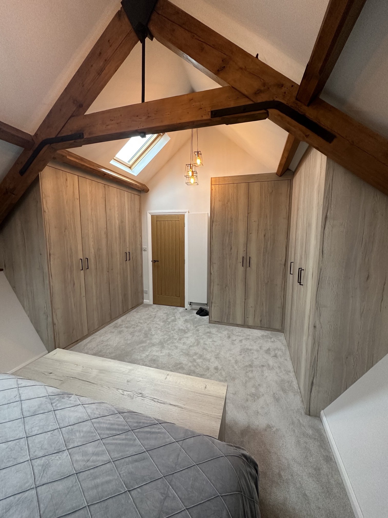 View of an attic room with wooden beams, built in wardrobes, a wooden door, a skylight, hanging lights, and a carpeted floor.