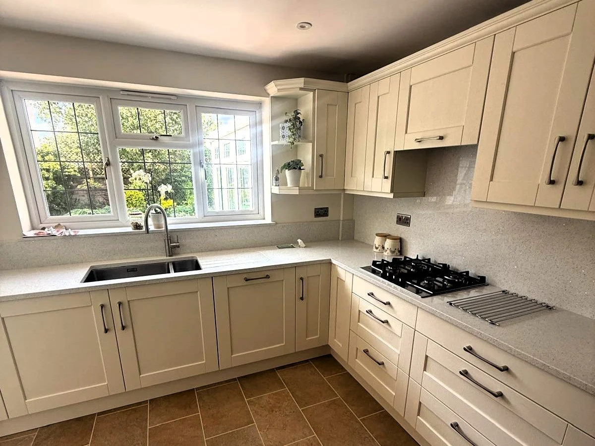 Modern kitchen with cream-colored cabinets, white countertop, black sink, and a window above the sink overlooking greenery.