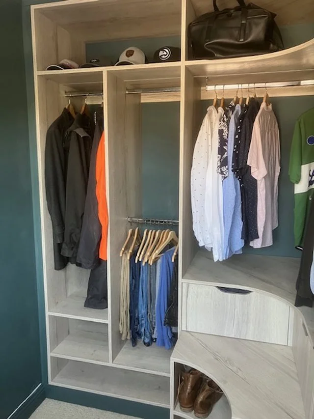 Open closet with hanging shirts, jackets, and trousers, a shelf with hats and a bag, and a shelf with boots at the bottom.