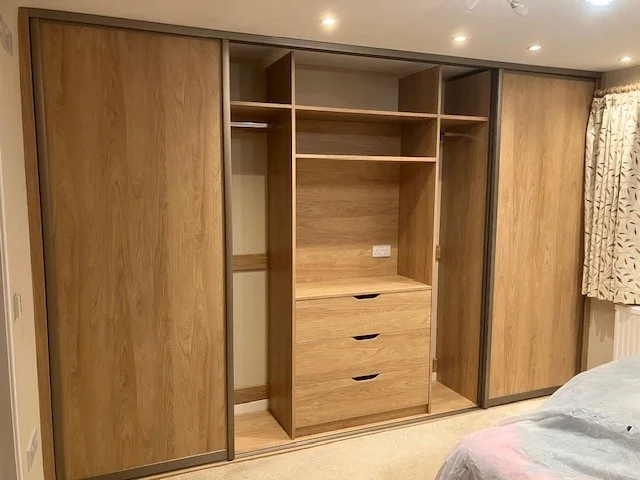 Wooden wardrobe with sliding doors, open to reveal shelves and drawers, in a bedroom with beige walls and curtains.