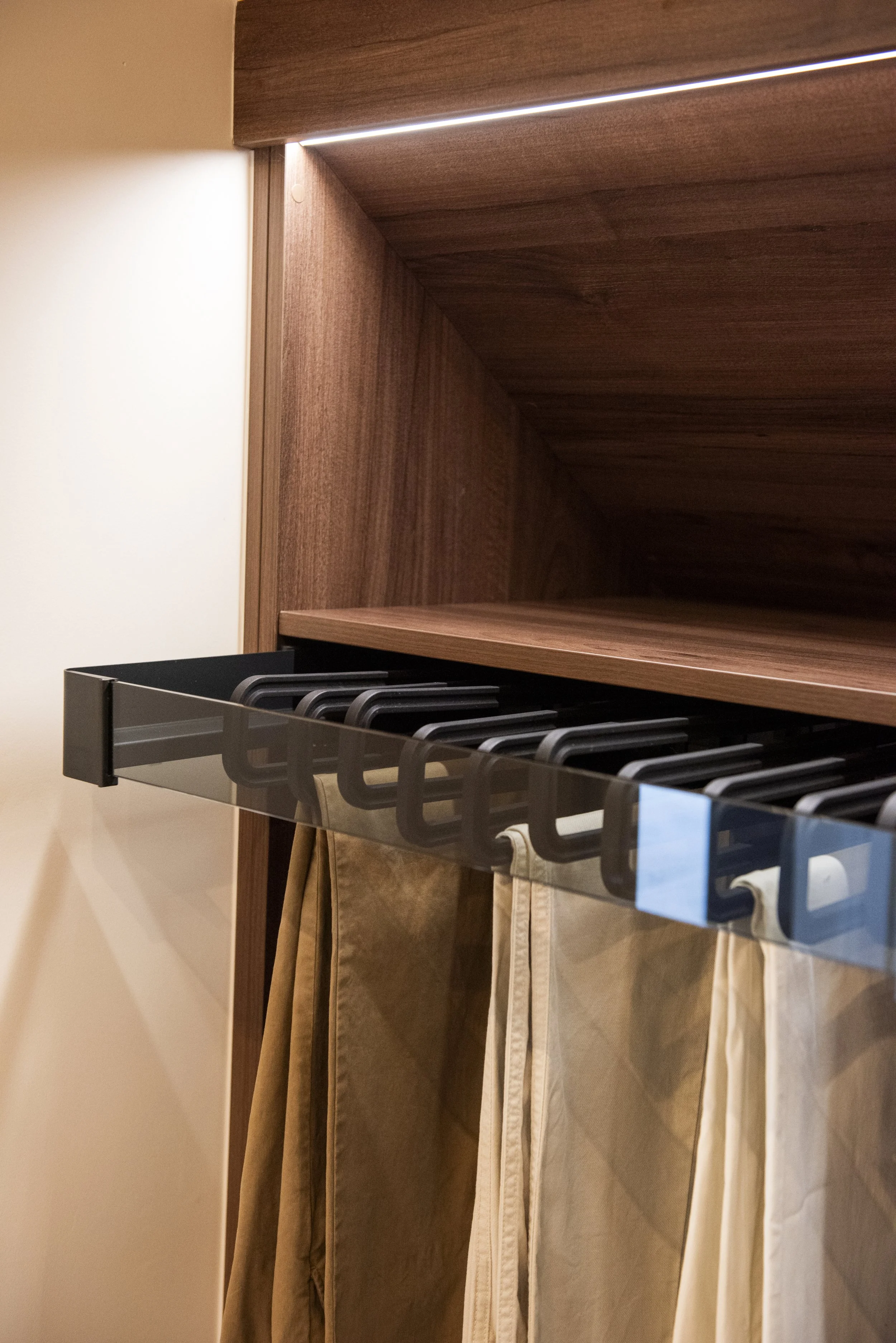 A close-up of a walk in wardrobe with a strip of LED lighting underneath, mounted above trouser hanging rail.