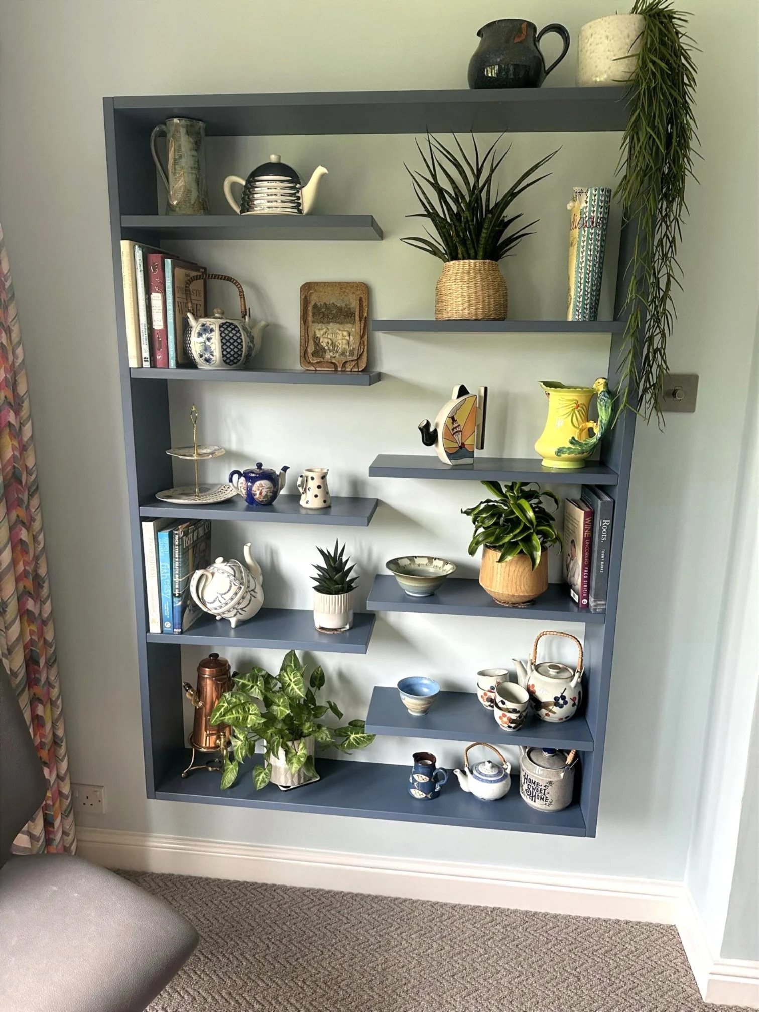 Gray bookshelf filled with decorative items, books, and potted plants against a light blue wall, with a beige curtain on the left and a brown carpet on the floor.