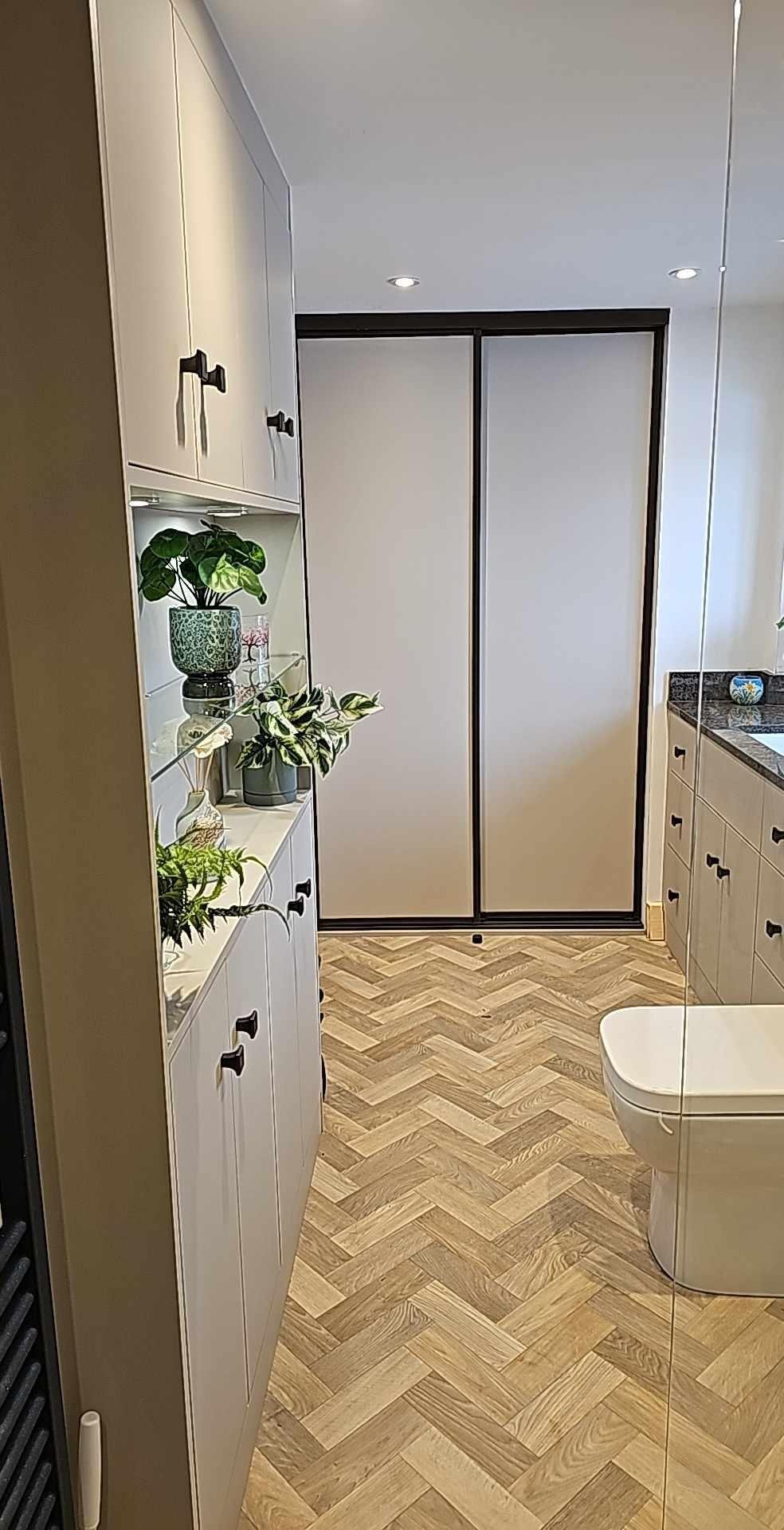 Interior of a modern bathroom with wooden herringbone flooring, white cabinetry, green plants, and a sliding door in the background.