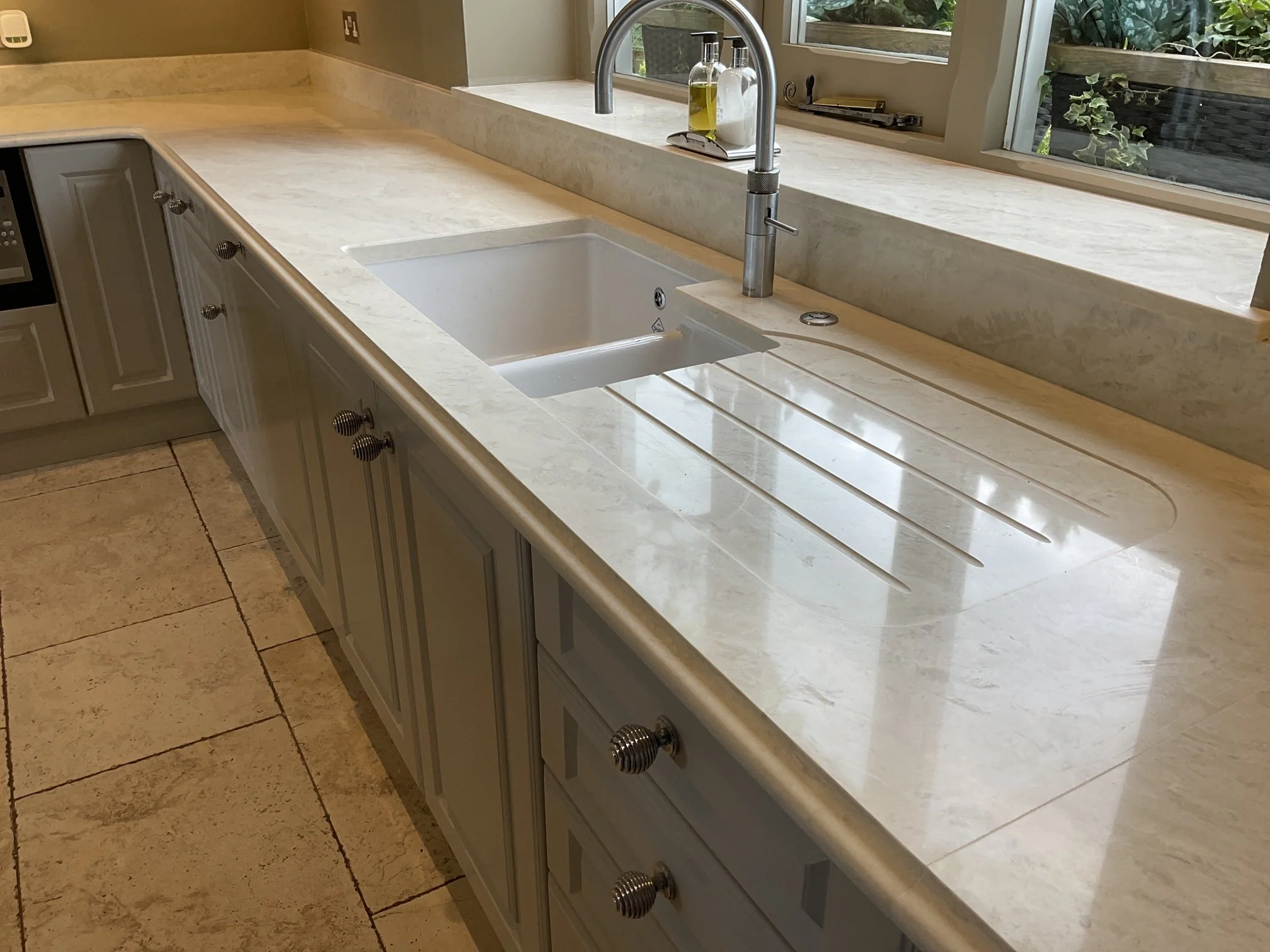 A kitchen countertop with a white sink, a modern silver faucet, a window with greenery outside.