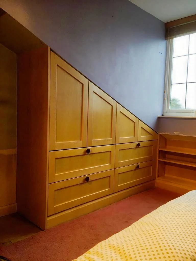 Wooden built-in storage unit under a sloped ceiling near a window in a bedroom