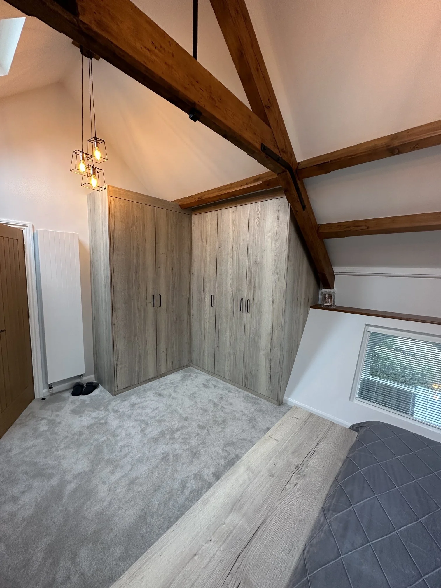 Interior of a room with vaulted ceiling and wooden beams, a large wooden built-in wardrobe, a carpeted floor, and a partial view of a bed.