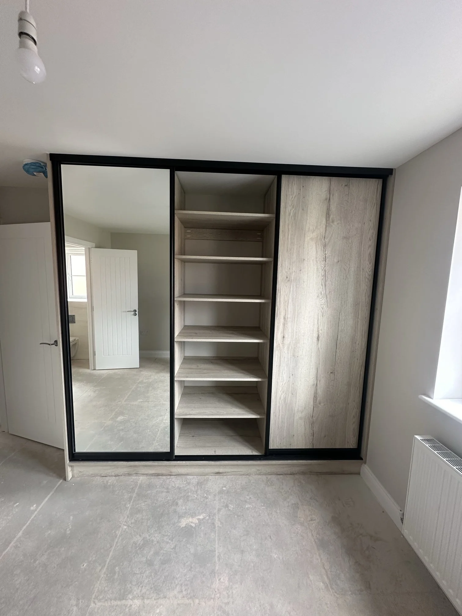 View of a partially open wardrobe with a sliding door mirror, open shelving, and a wooden panel. 