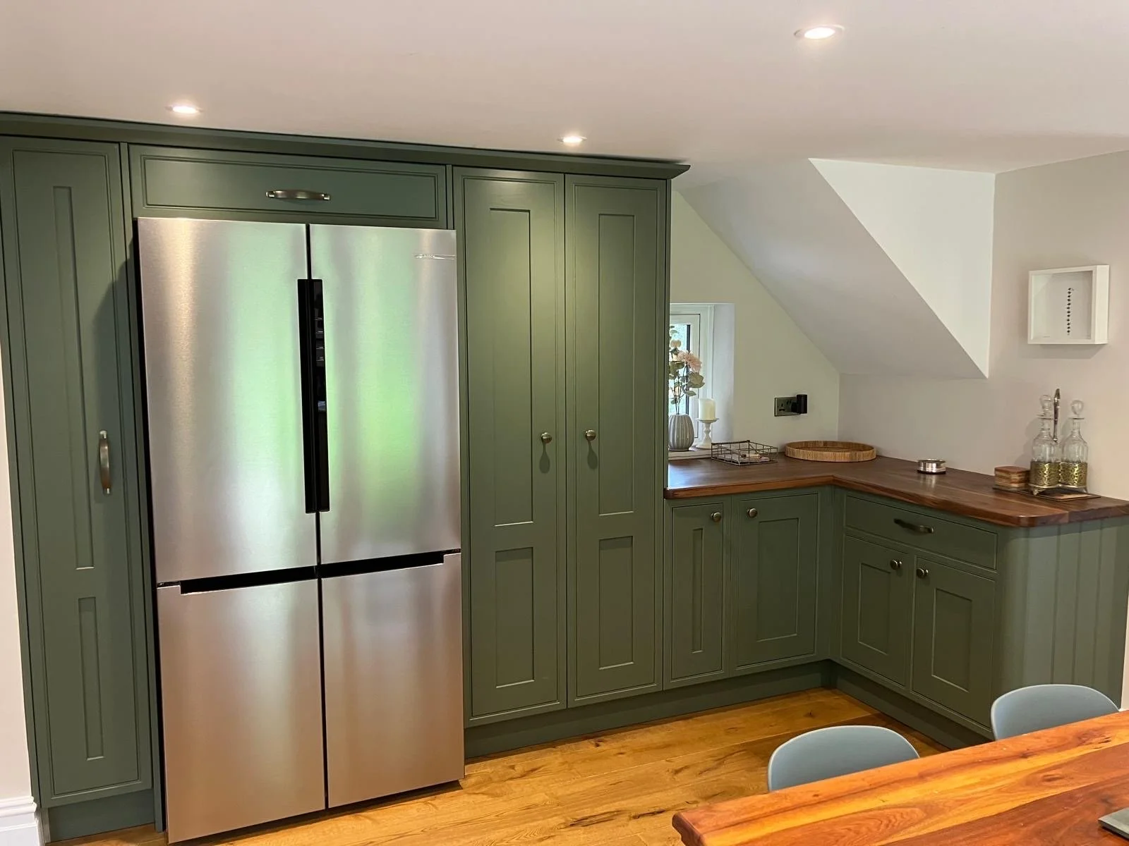 Green kitchen cabinetry with a stainless steel refrigerator, a small window with a vase and candles, and a wooden countertop with glass bottles and decorative items.