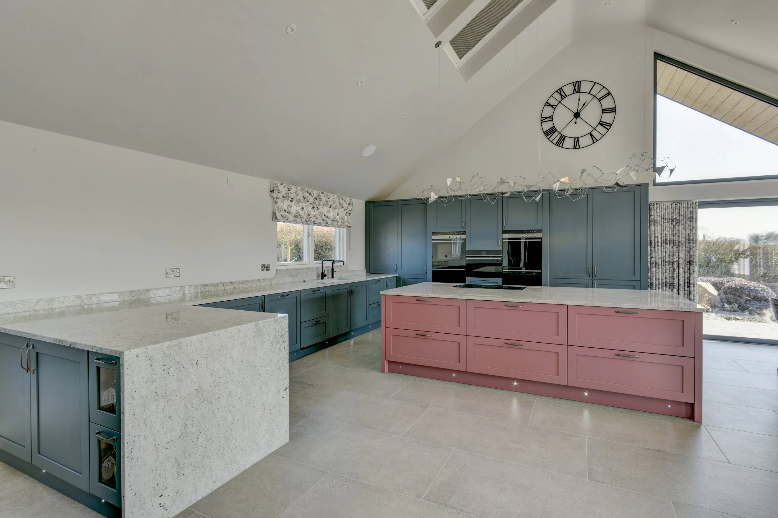 Large contemporary kitchen with blue cabinetry, pink island, white grantie countertops, large window, and high ceiling with clock.