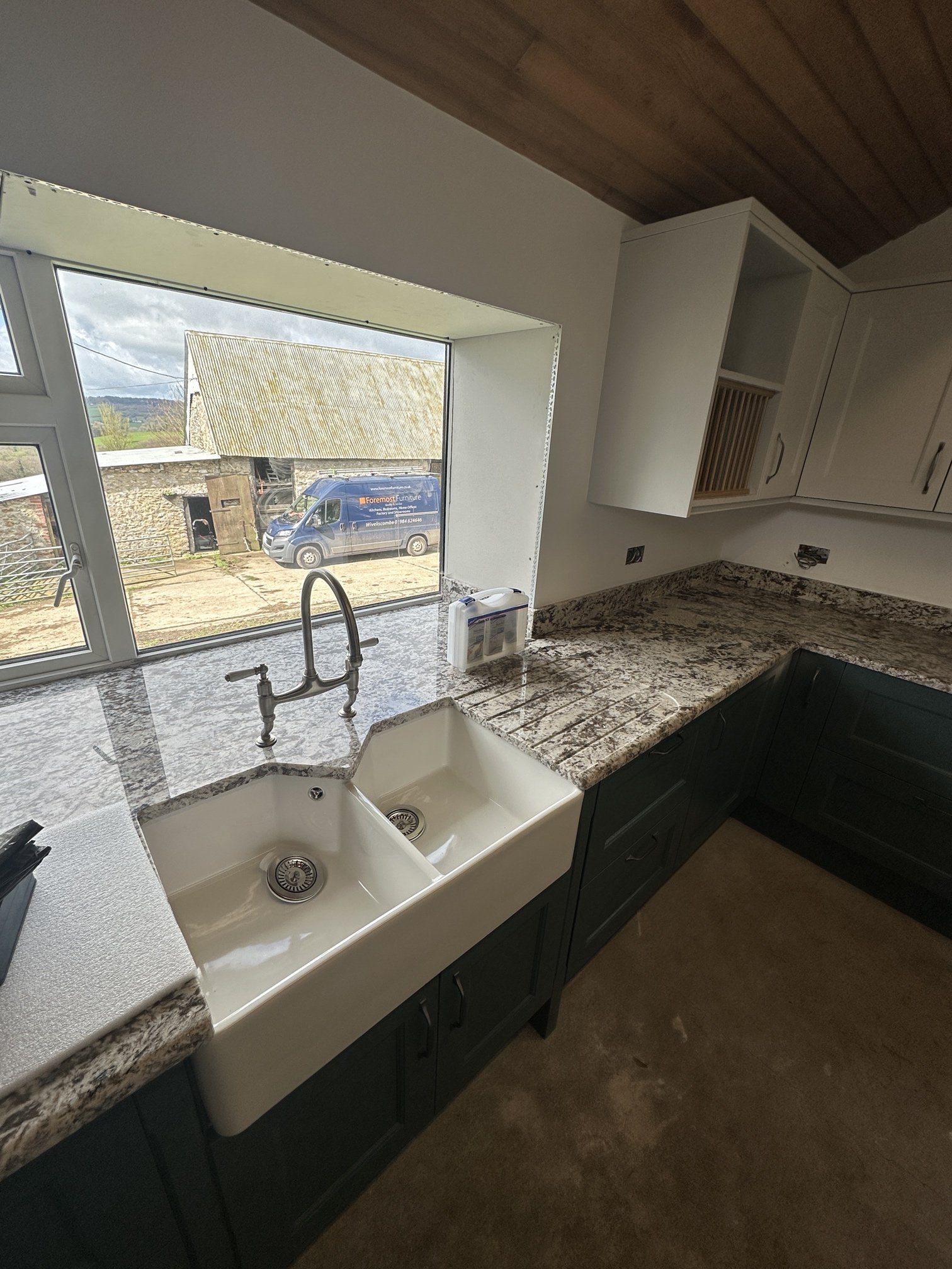 Kitchen with double farmhouse sink, granite countertops, white upper cabinets, and dark lower cabinets. There is a large window above the sink showing an outdoor scene with a van, a building, and cloudy sky.