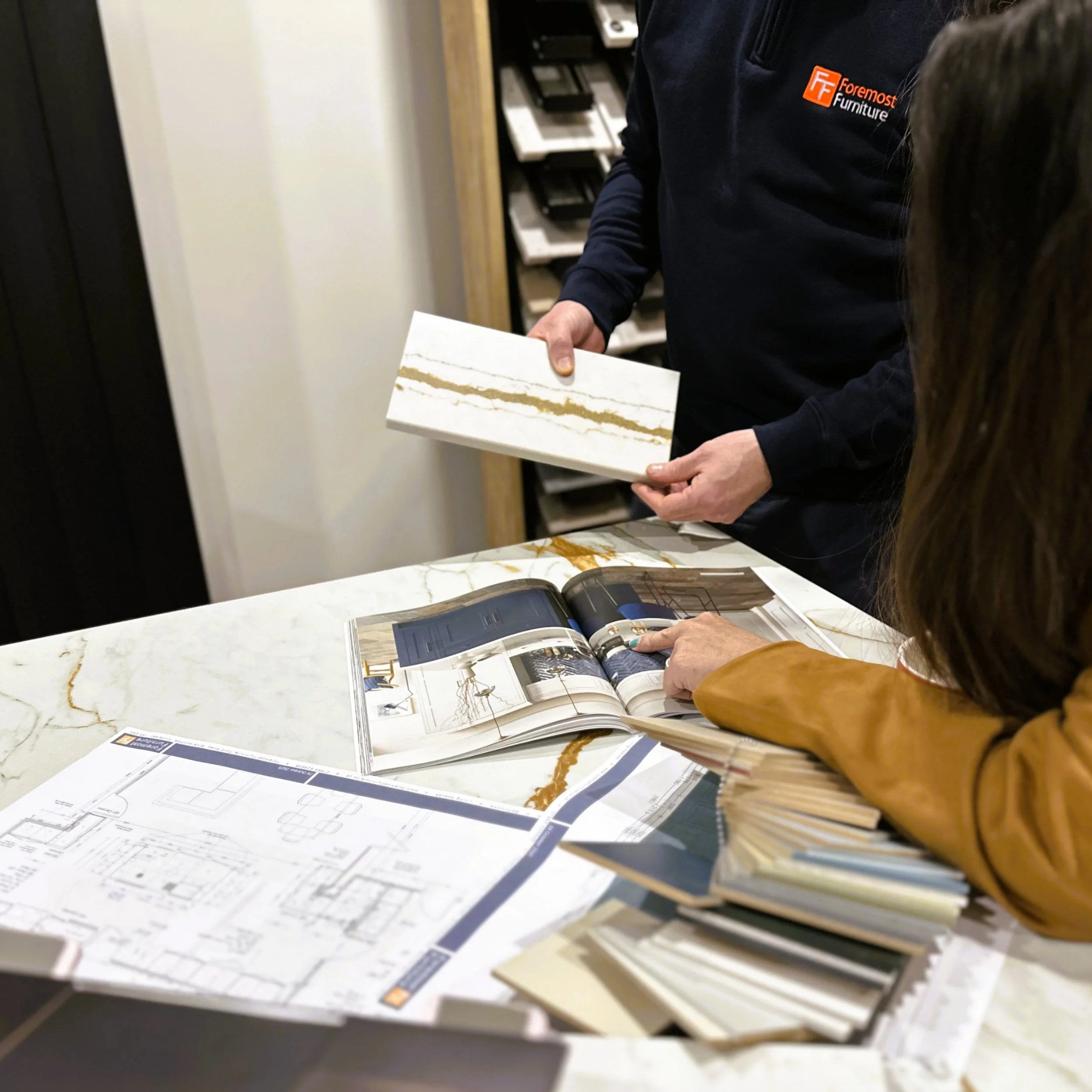 Employee holding a sample in a store and a customer pointing to an open catalog page on granite countertop.