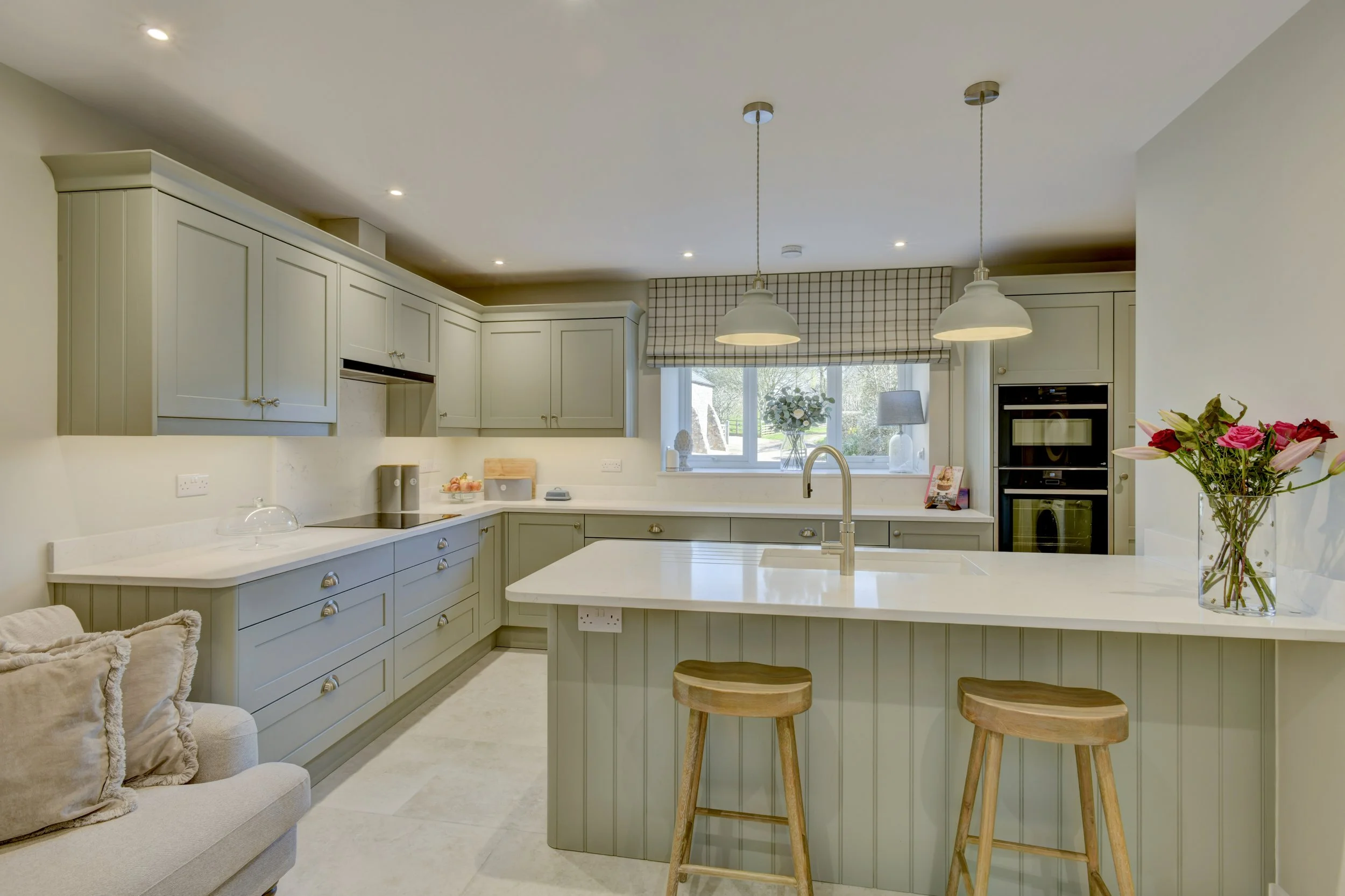 A contemporary kitchen with light grey cabinets, white Quartz countertops, two pendant lights hanging over the counter, a window with a checkered valance, a vase of flowers, and a breakfast bar with two wooden stools.