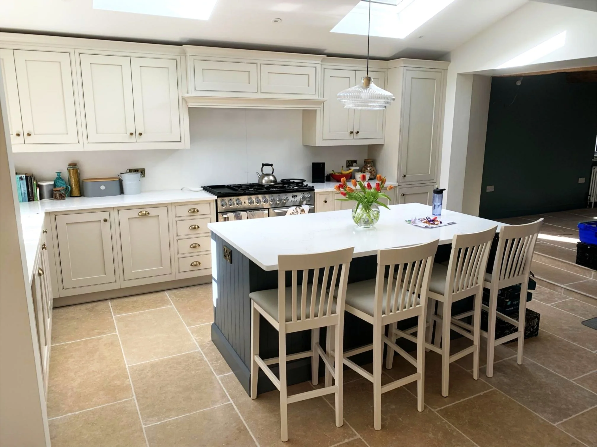 A traditional kitchen with white cabinetry, a white island, and a grey island with stools. The kitchen has a range cooker and various kitchen items. The room features natural light from skylights.