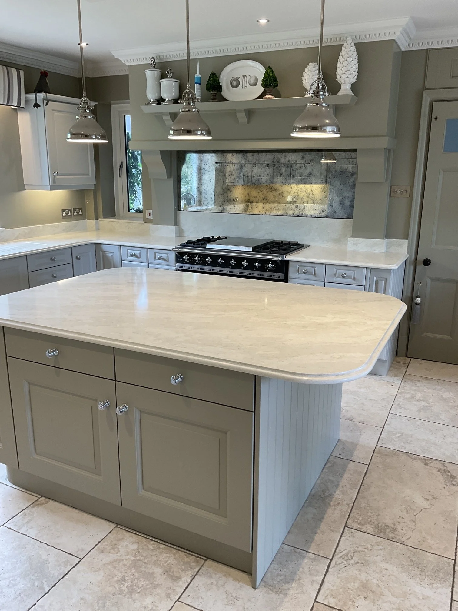 A kitchen with a central island featuring a light-colored countertop and cabinets with silver knobs. The background includes a stove with a reflective backsplash, white cabinetry, and a window. 
