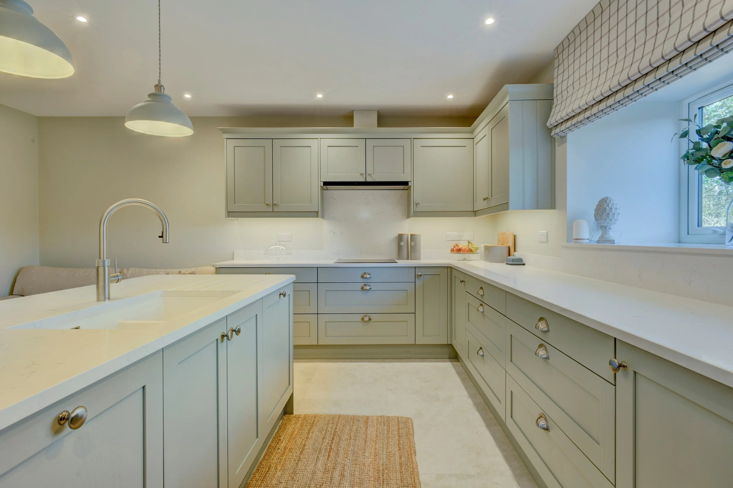A contemporary kitchen with greige cabinets, Quartz countertops, a large window with a checkered Roman shade, and a beige rug on the floor.