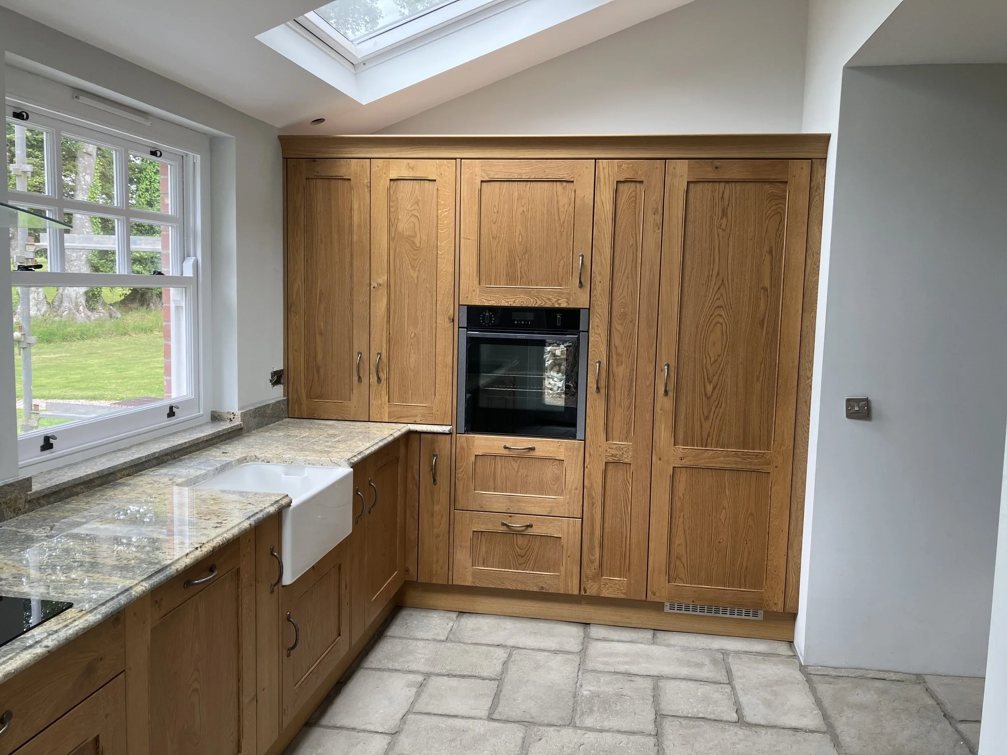Kitchen with wooden cabinets, granite countertops, a white farmhouse sink, a built-in oven, and a large window with a view of green trees outside. Skylight above provides natural light.