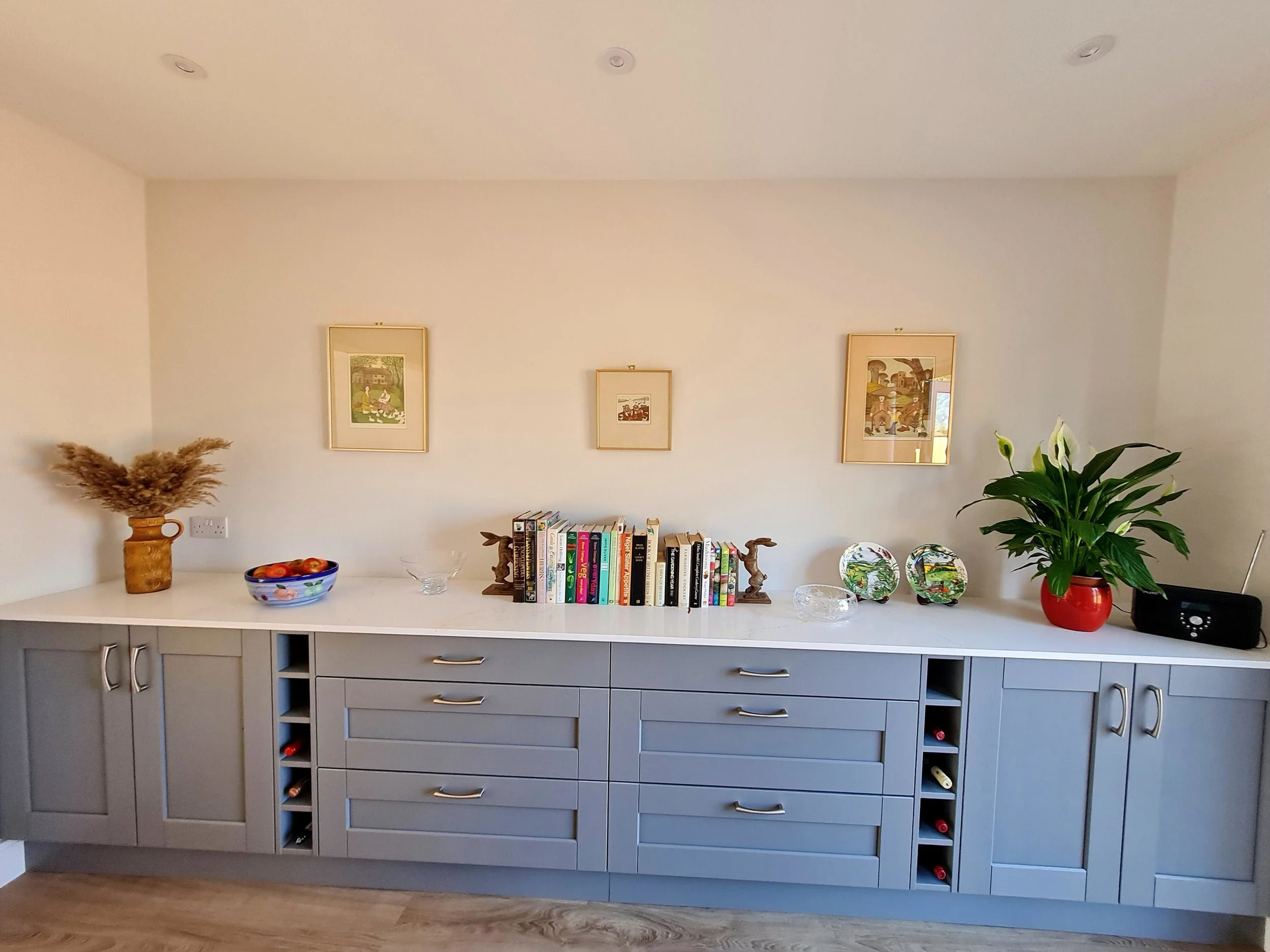 A light blue kitchen cabinet with multiple drawers and wine bottle slots, decorated with a tall orange vase with dried plants, a bowl of apples, various books, decorative plates, and a potted plant, against a white wall with three framed pictures.