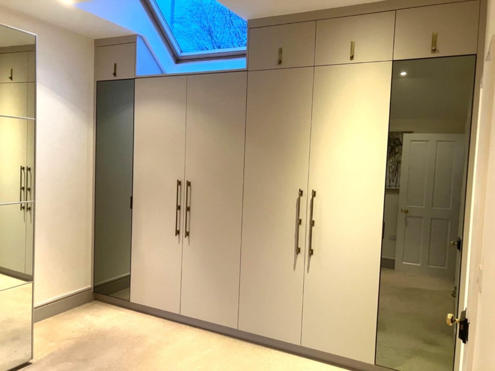 A room with built-in beige wardrobes with brass handles, a mirror on the left, and a skylight window showing blue sky and tree branches outside.