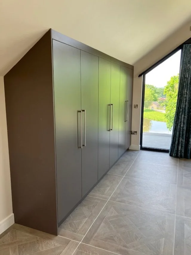Grey built-in wardrobe t with long vertical handles in a room with large window and tile flooring.