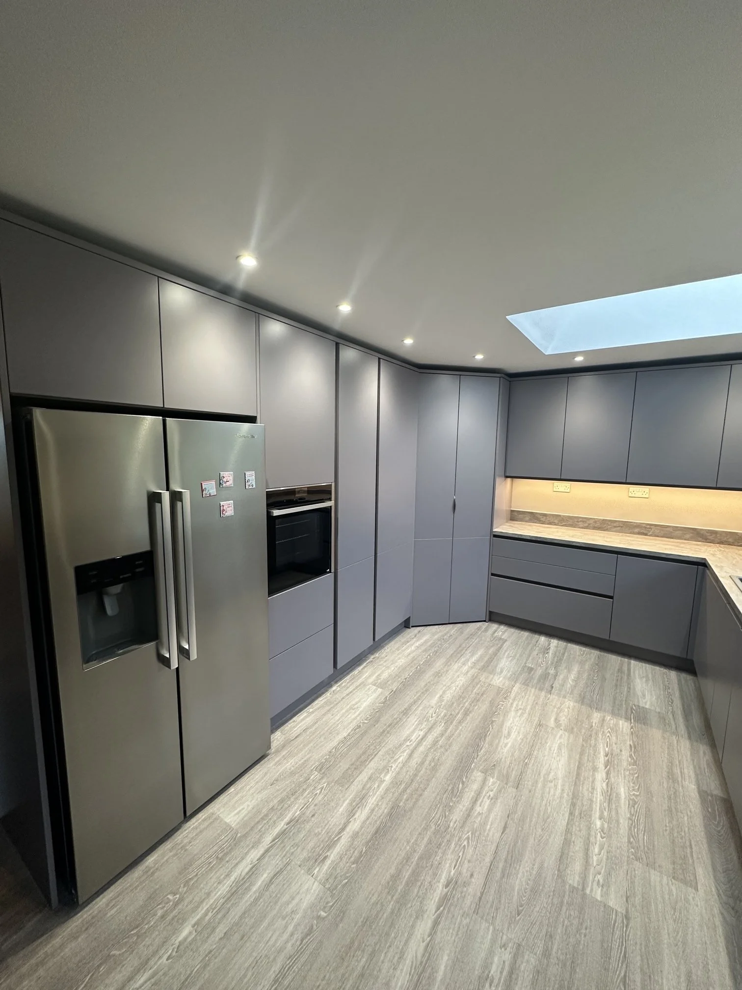 Modern kitchen with grey cabinets, stainless steel refrigerator, built-in oven, and skylight illuminating the space.