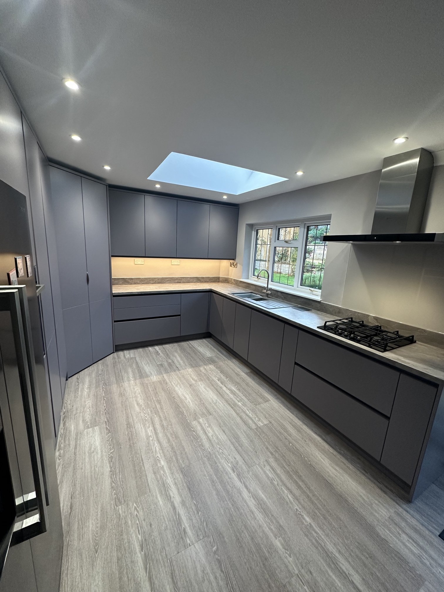 Modern kitchen with grey cabinets, marble countertops, wooden flooring, a skylight, and a window overlooking a garden.