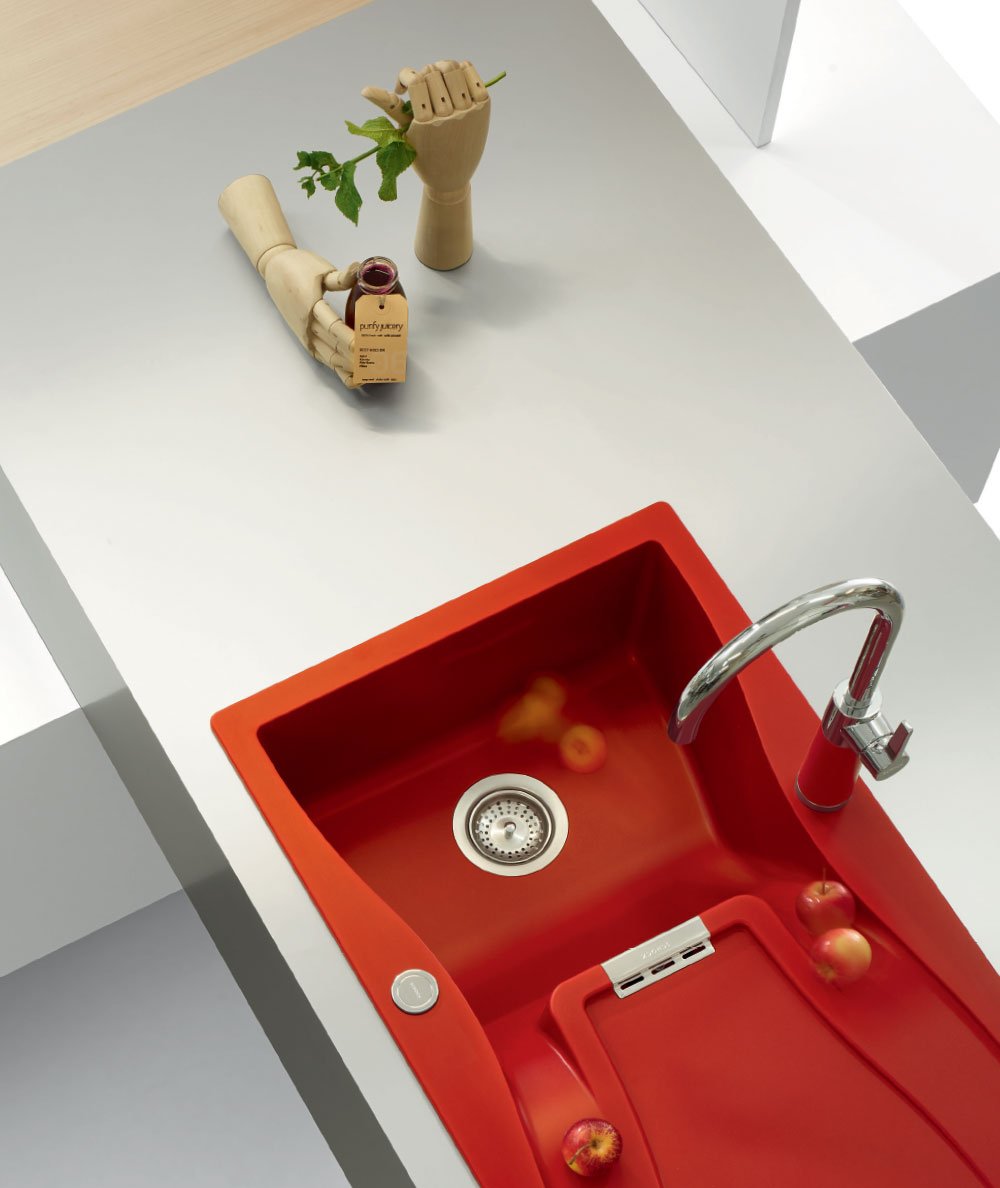 A kitchen countertop with a Schock red sink, apples, a wooden mannequin hand holding a green leaf, and a bottle of jam.