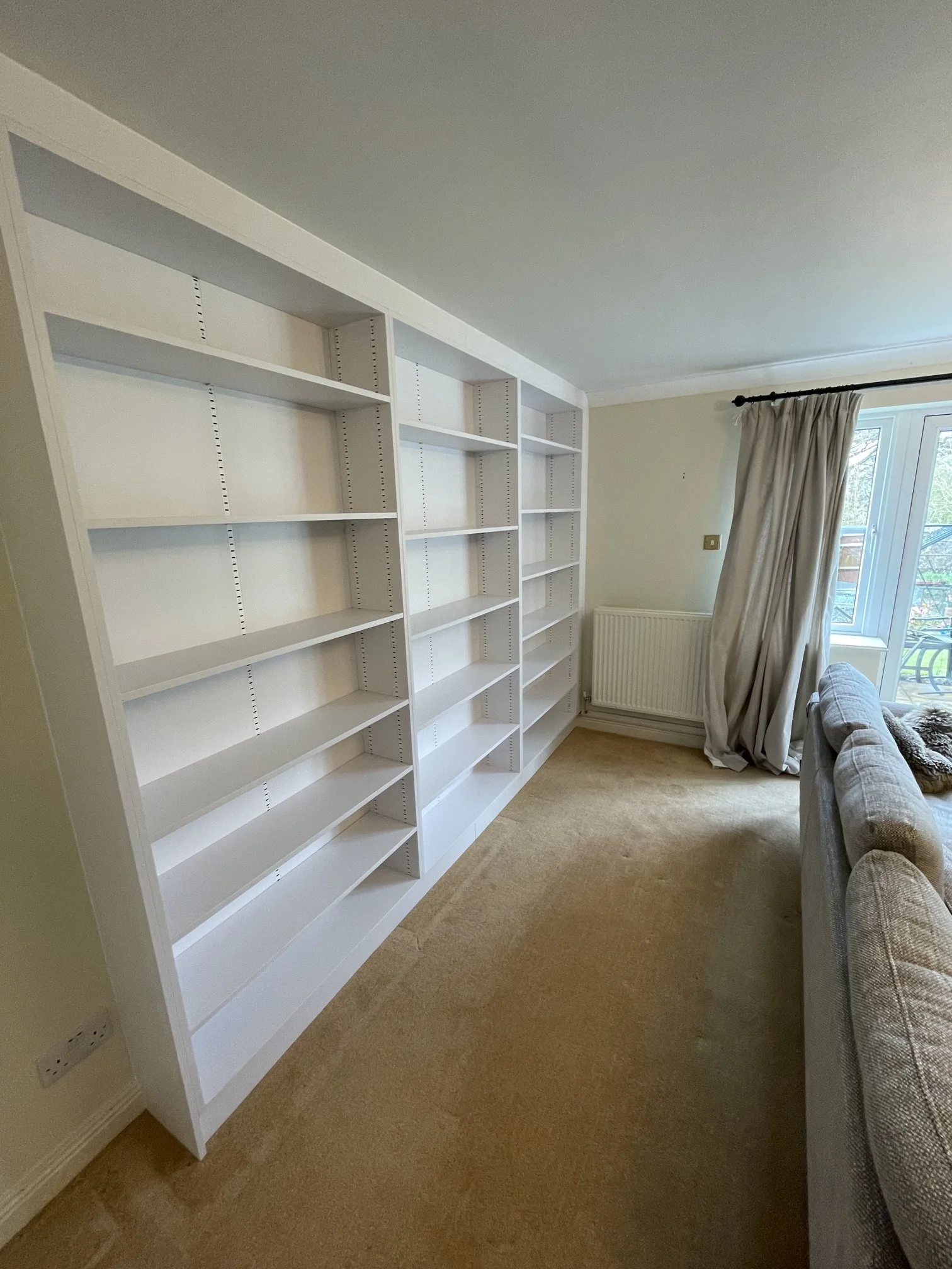 Office with white empty bookshelves, a window with beige curtains, a beige carpet, and a patterned sofa.