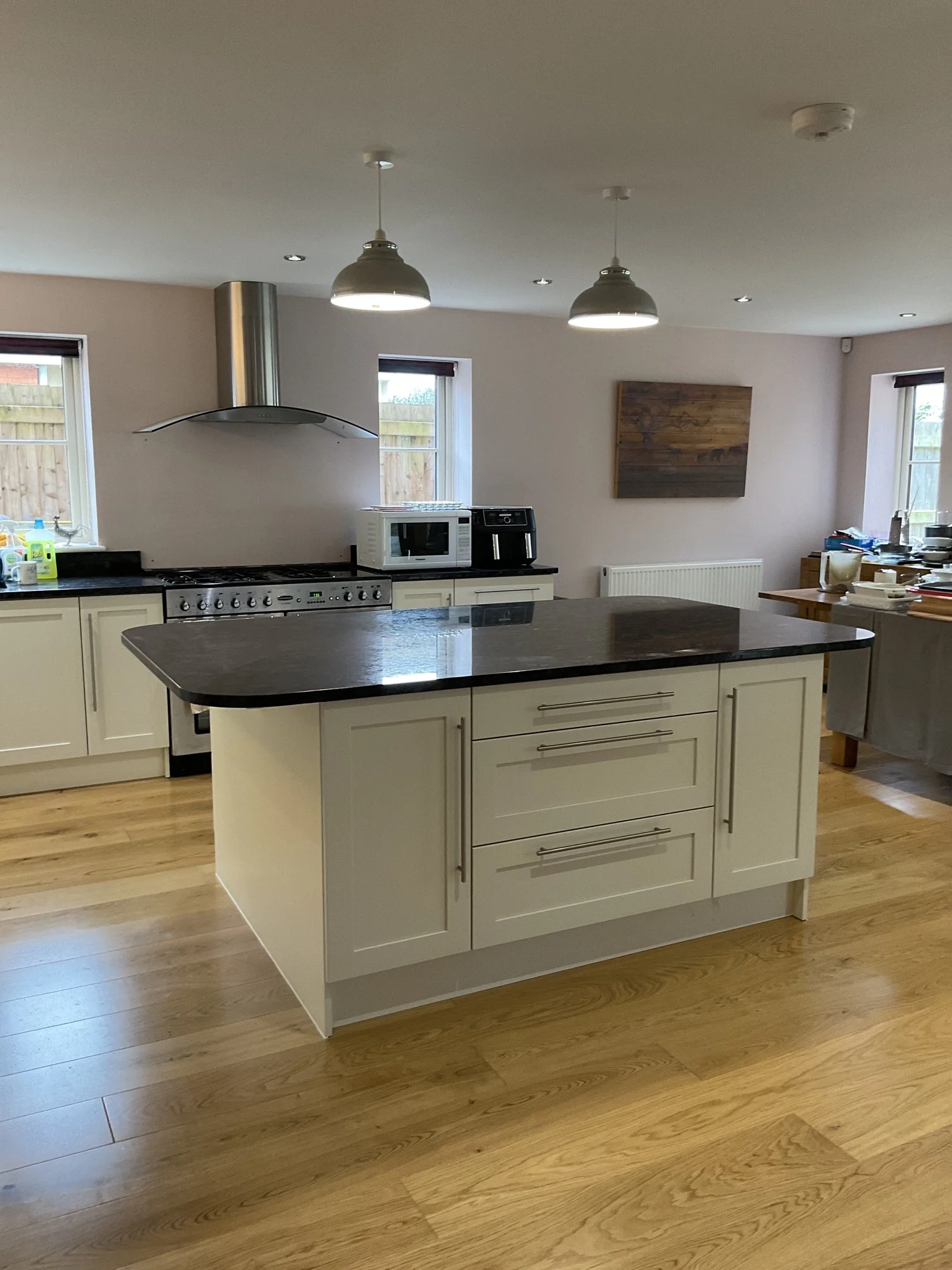Kitchen with a central island featuring a dark countertop, white cabinets with silver handles, a stove with oven, microwave, coffee machine, two windows with blinds, hanging pendant lights, a wooden wall art, and various kitchen items on the counters