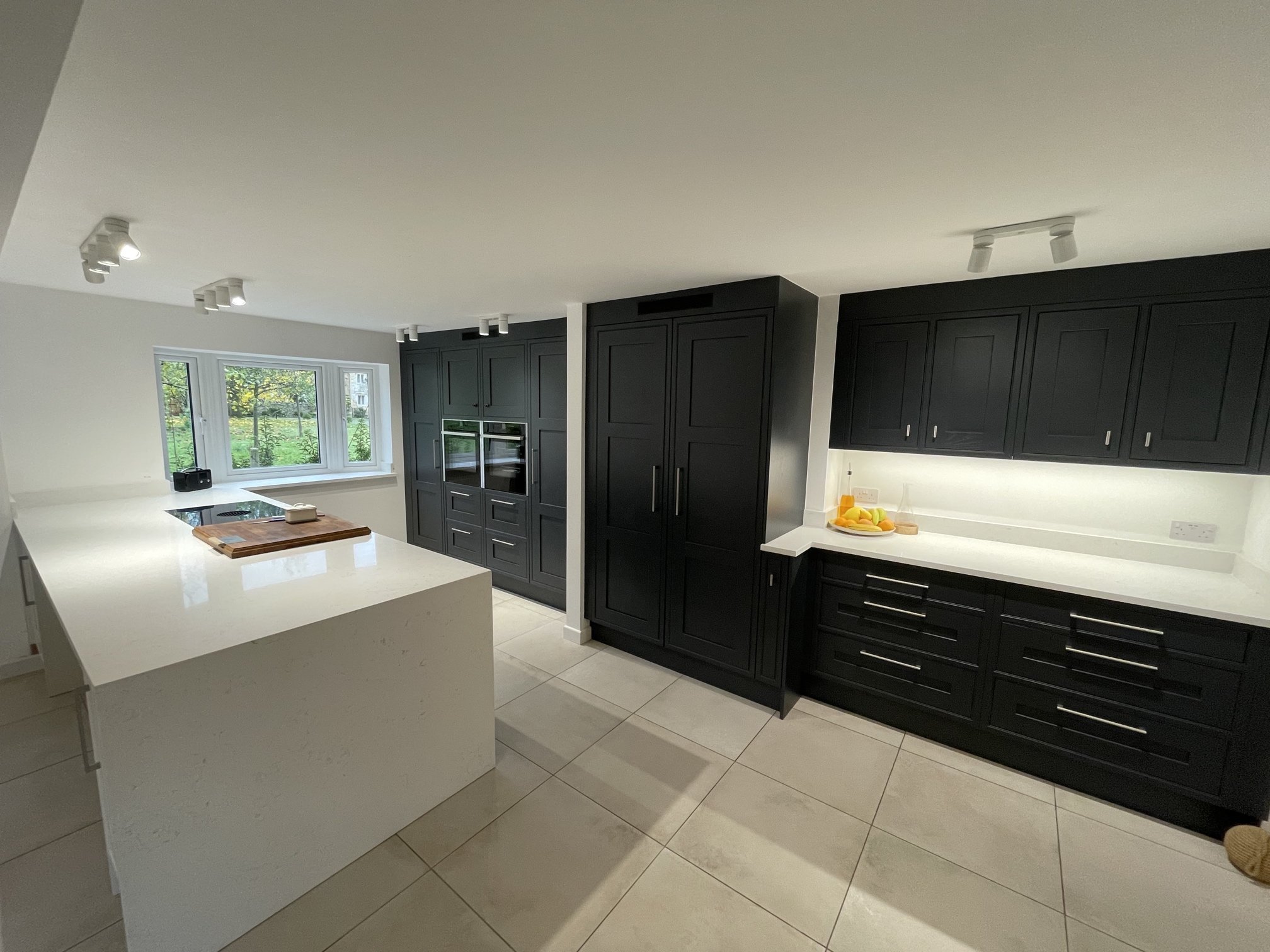 Modern kitchen with white countertops, black cabinets, a large window, and white tiled flooring.