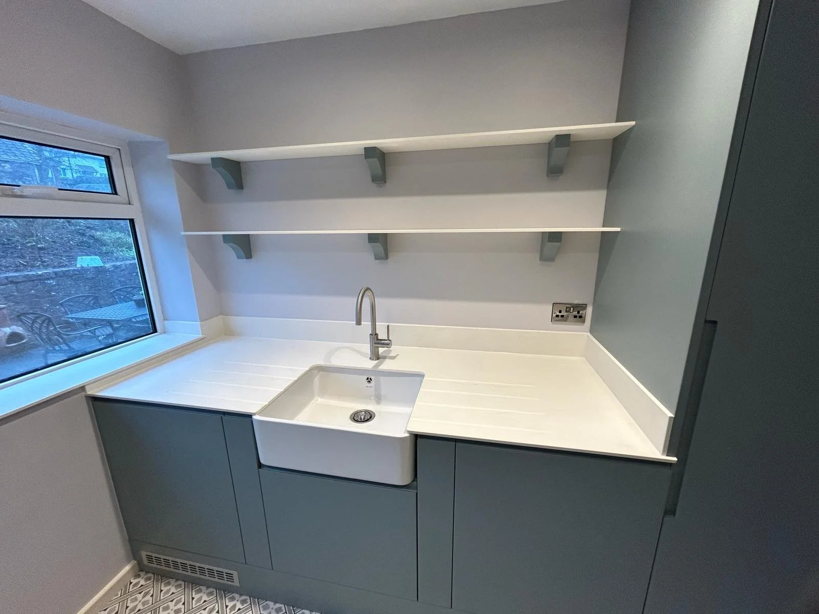 Utility room with a white farmhouse sink, light grey cabinets, open white shelves with grey brackets, a window showing an outdoor seating area.
